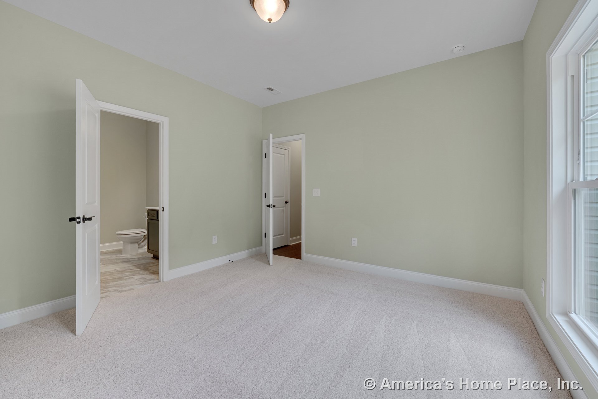 Bedroom with light carpet flooring, soft green walls, white trim and baseboards, double white paneled doors including access to en suite bathroom, large window with white casing