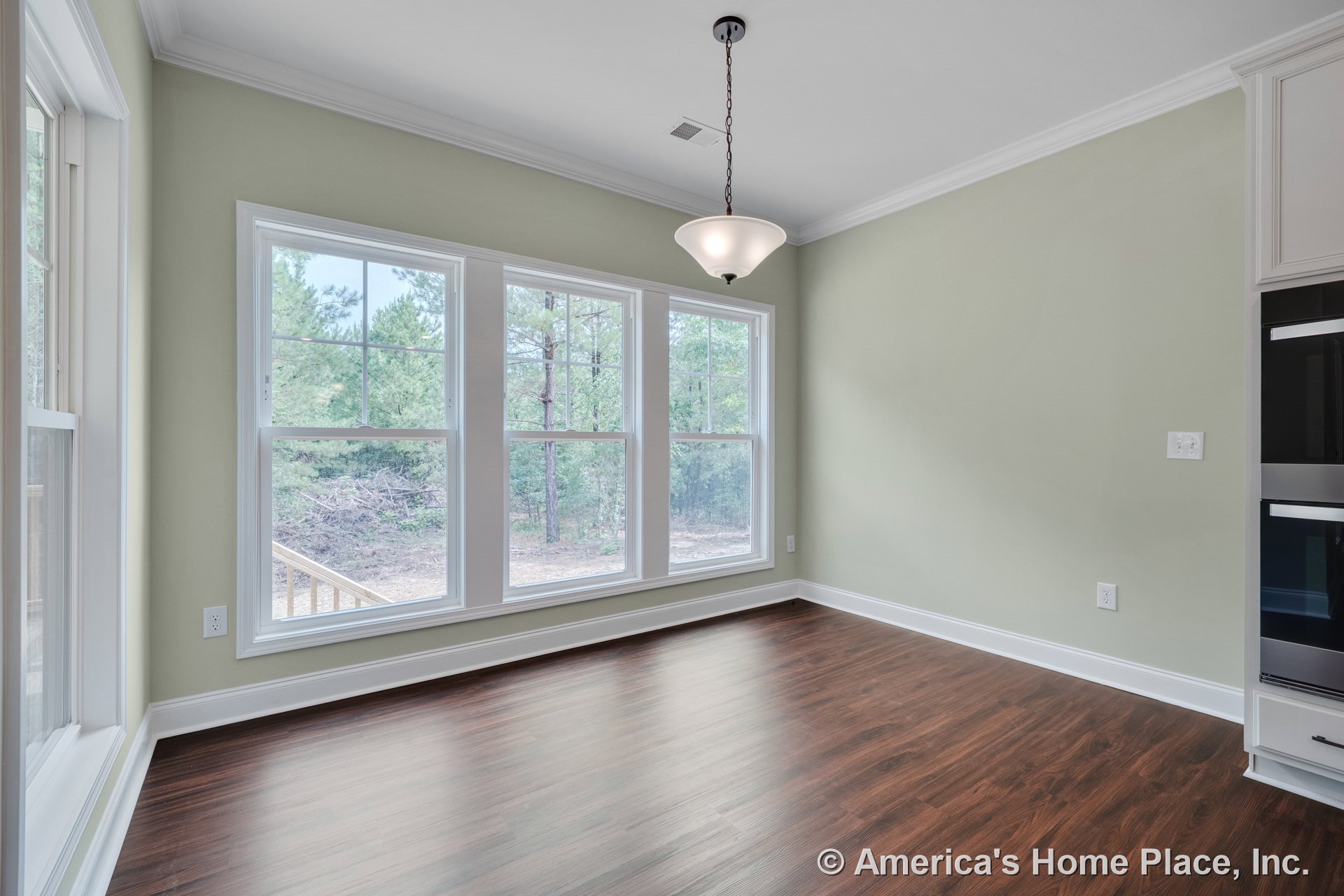 Dining area with triple window wall, white crown molding, wood-look plank flooring, soft green painted walls, pendant ceiling light, baseboard trim, and built-in cabinetry edge.