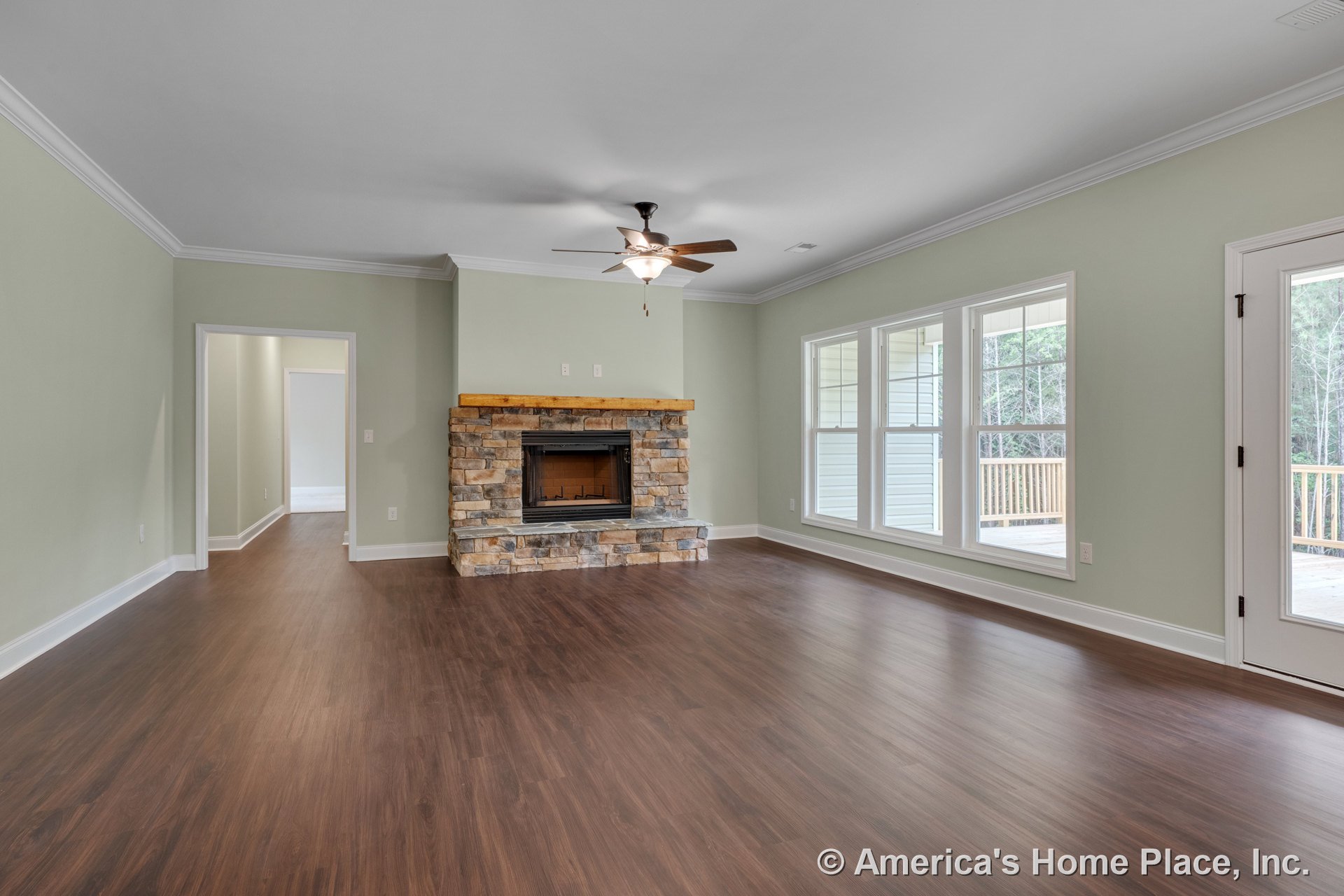 Stone fireplace with wood mantel, large windows facing porch, crown molding, ceiling fan with light, wood-look flooring, and baseboards in spacious living room.