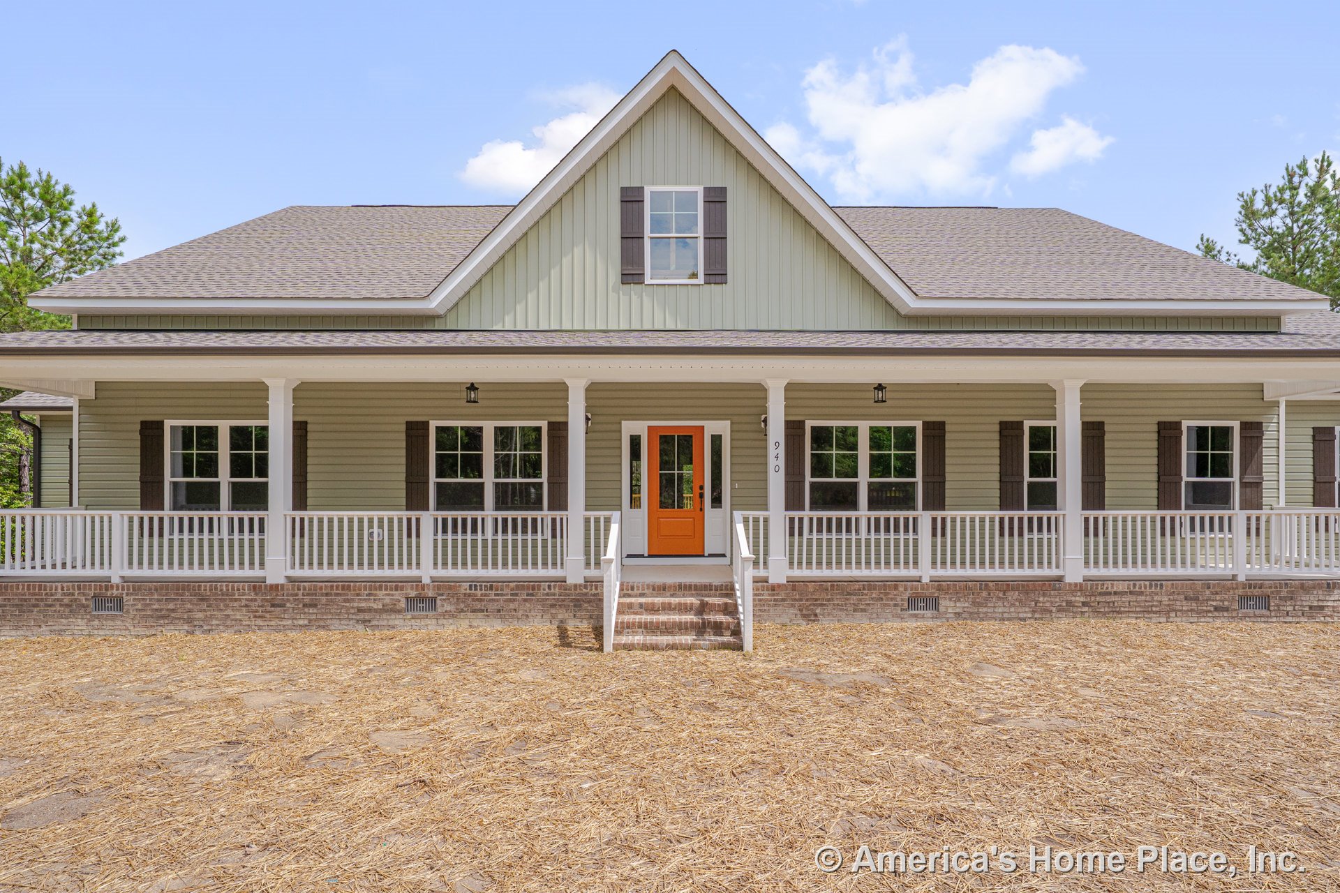 Large covered front porch with white railings, orange entry door, brick foundation, multiple double-hung windows with shutters, gable roof with shingles, white columns, exterior