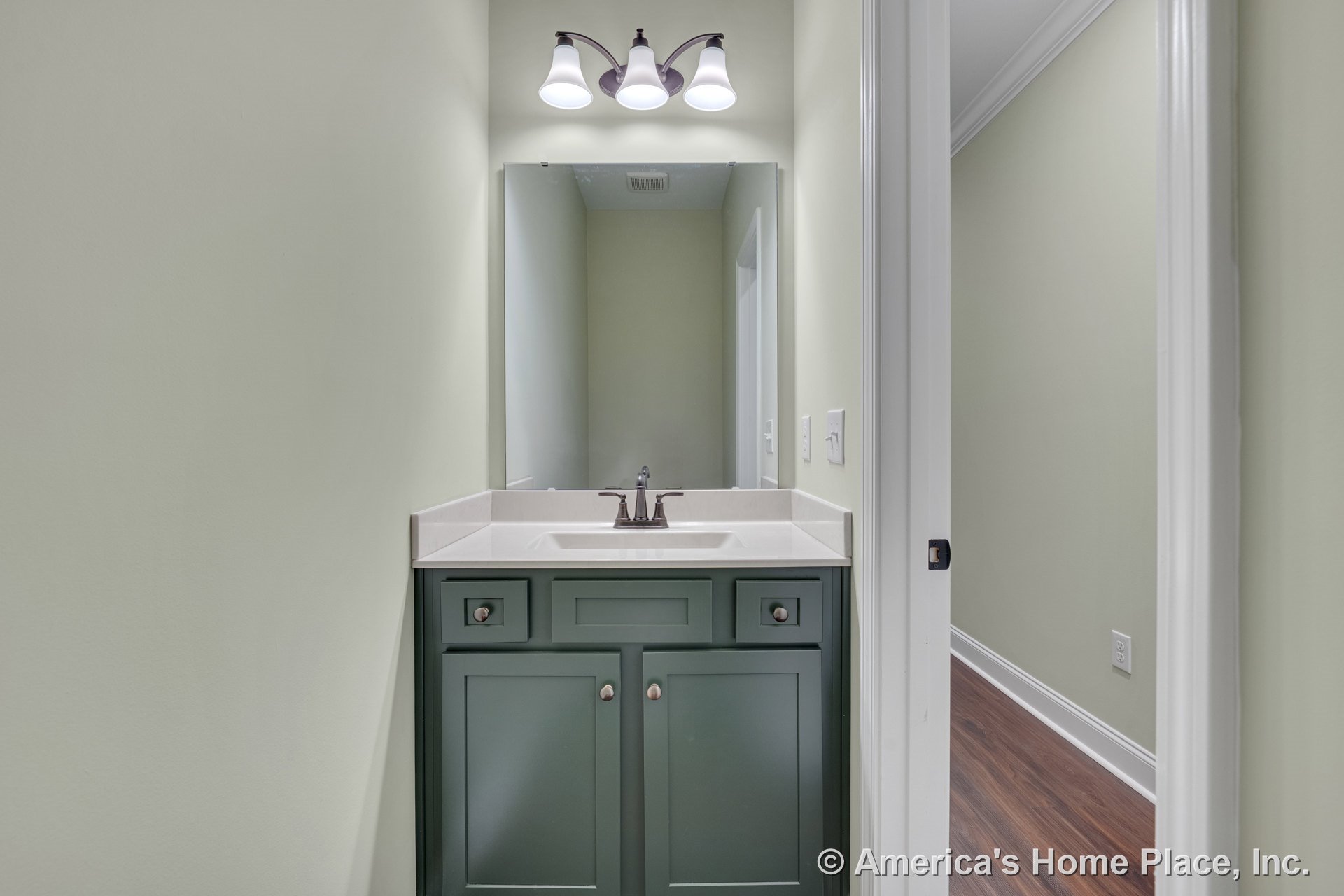 Green vanity cabinet with white solid surface countertop, rectangular wall-mounted mirror, three-light overhead fixture, light green painted walls, white trim, and door frame.