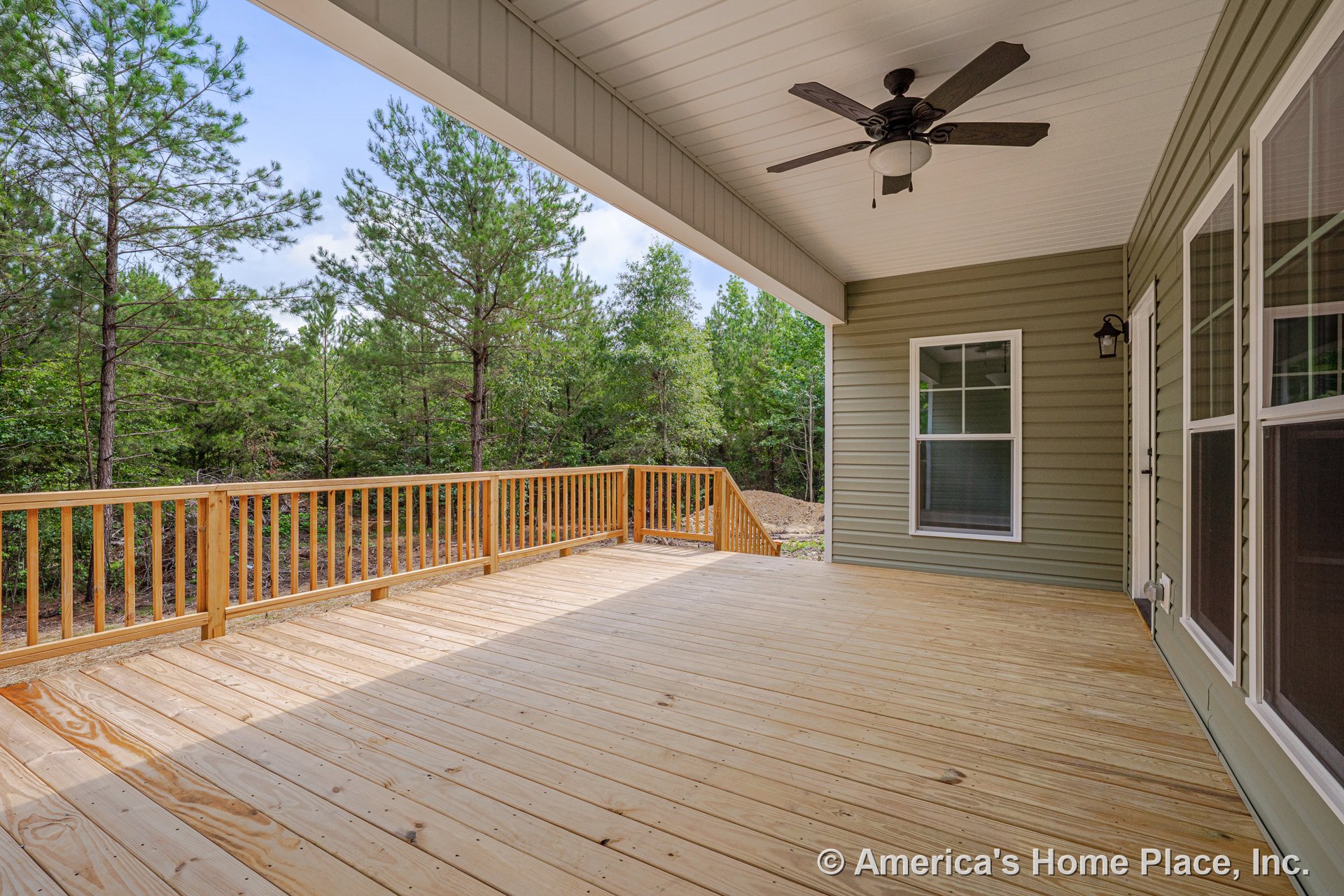 Covered wooden deck with ceiling fan, wood railing, green siding exterior, large windows, and wood flooring overlooking wooded landscape.
