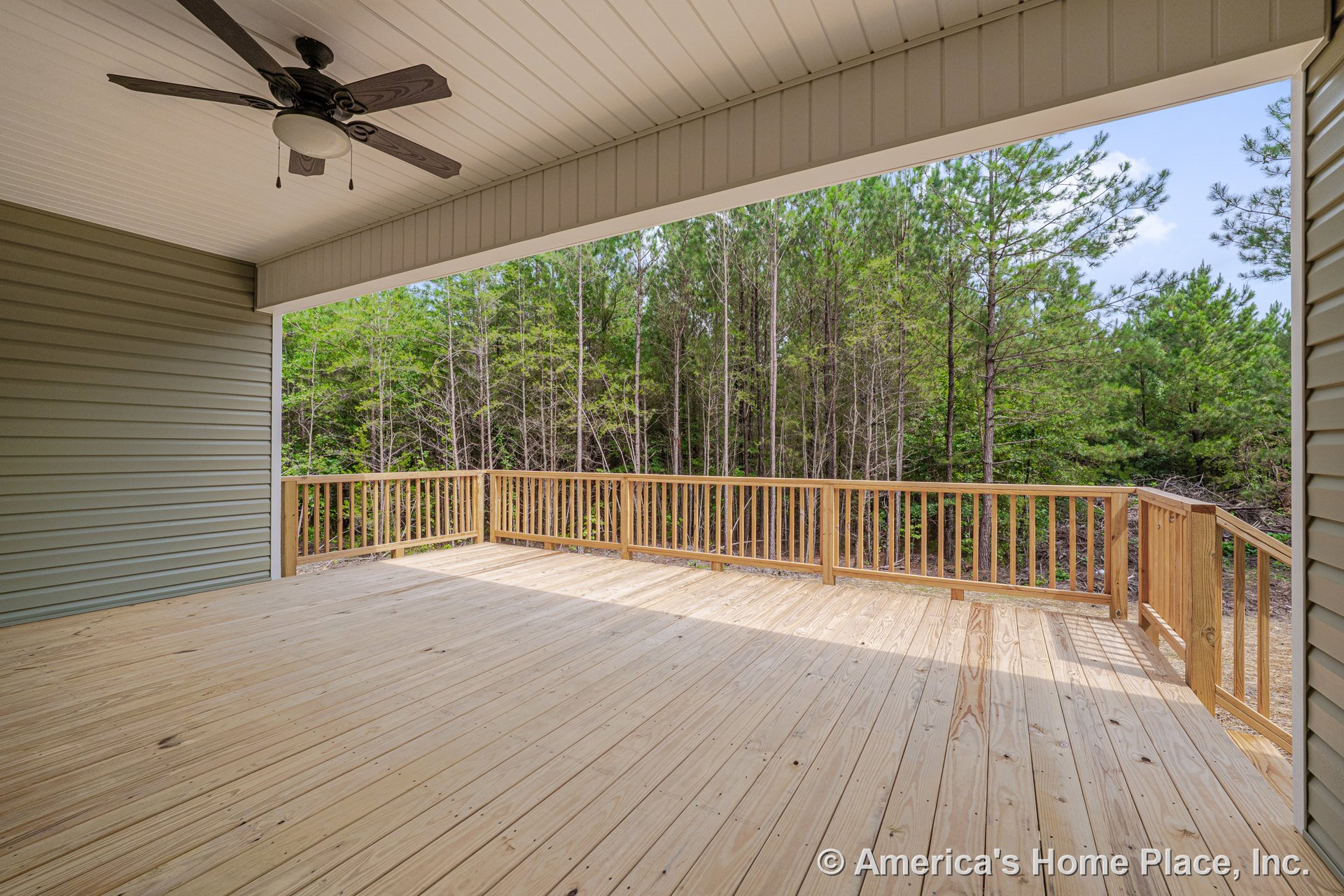 Covered wooden deck with plank flooring, ceiling fan, natural wood railing, exterior siding, and forest view beyond railing.