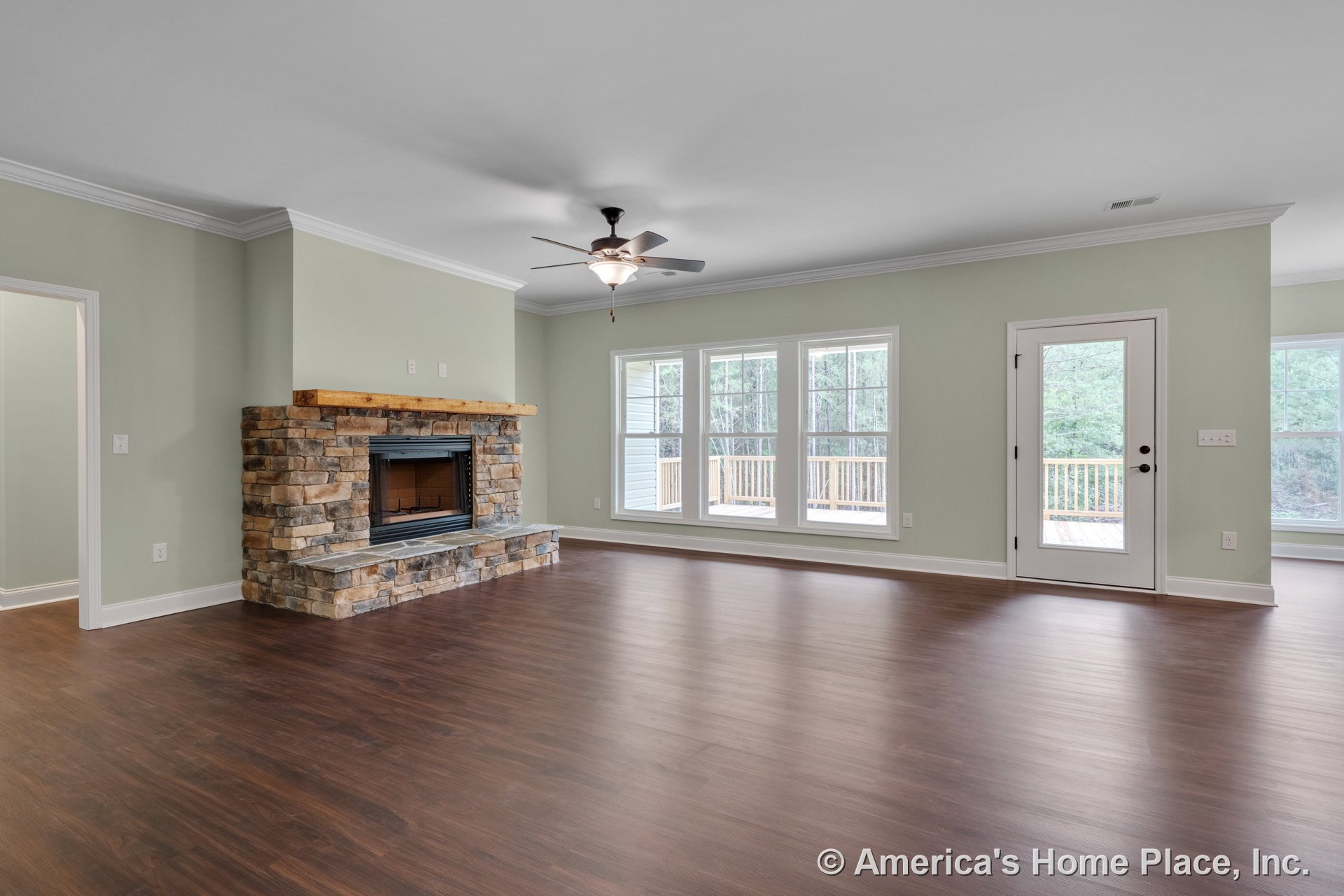 Stone fireplace with wood mantel, three large windows, wood-look flooring, ceiling fan with light fixture, crown molding trim, and glass exterior door.