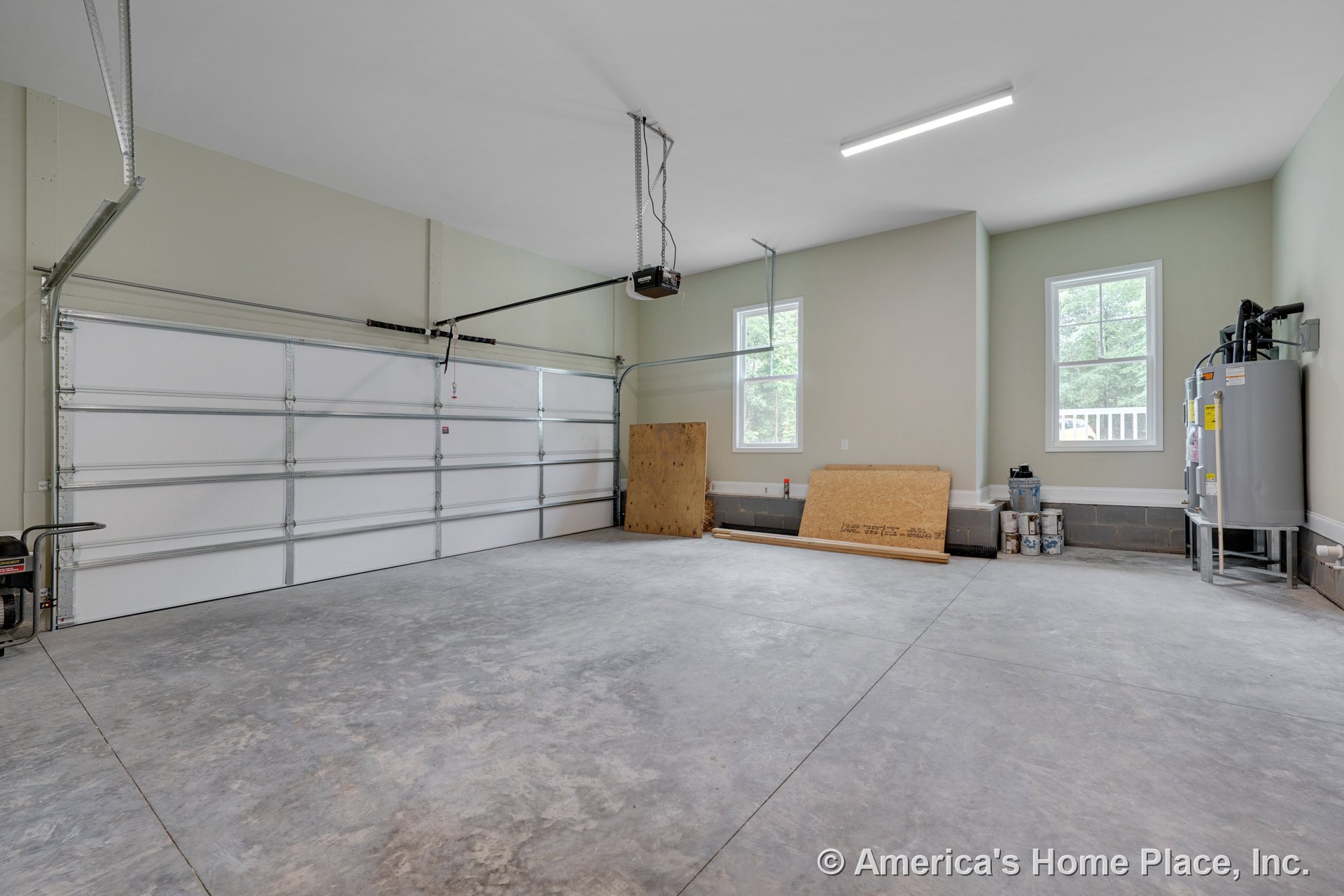 Double overhead garage door with concrete slab floor, two large windows providing natural light, wall-mounted water heater, fluorescent ceiling light, unfinished wall trim, and