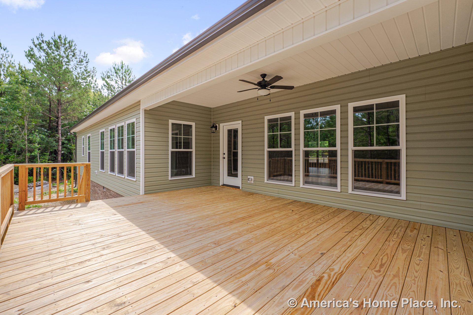 Covered wooden porch with green vinyl siding, white trim double-hung windows, ceiling fan mounted under roof, railing along edge, and single exterior door.