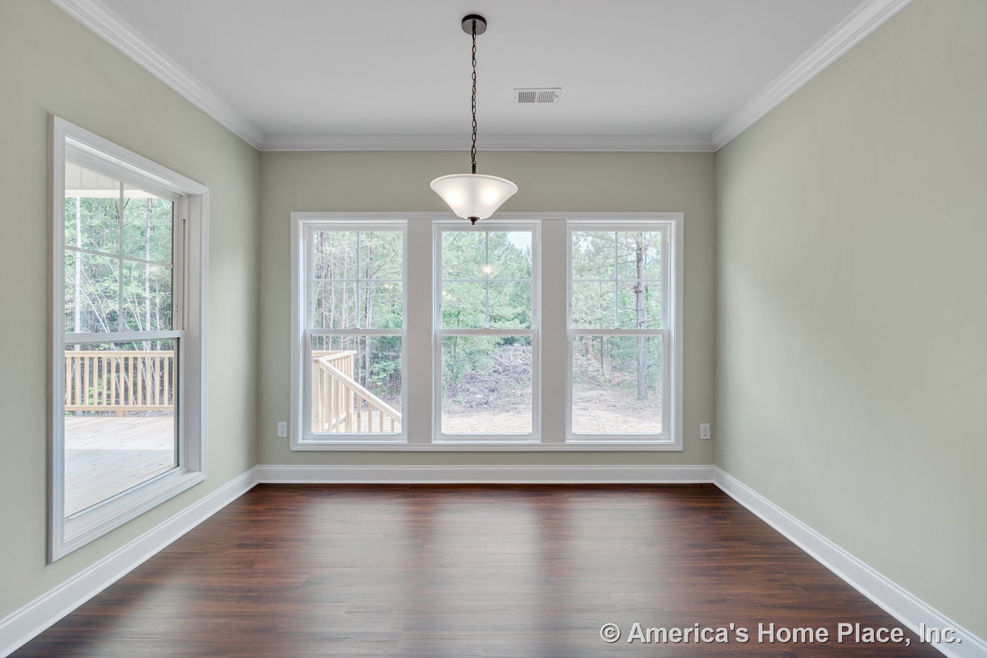 Dining area with triple window wall, wood-look flooring, white crown molding, central pendant light, soft green walls, baseboards, ceiling vent, and porch visible through windows.