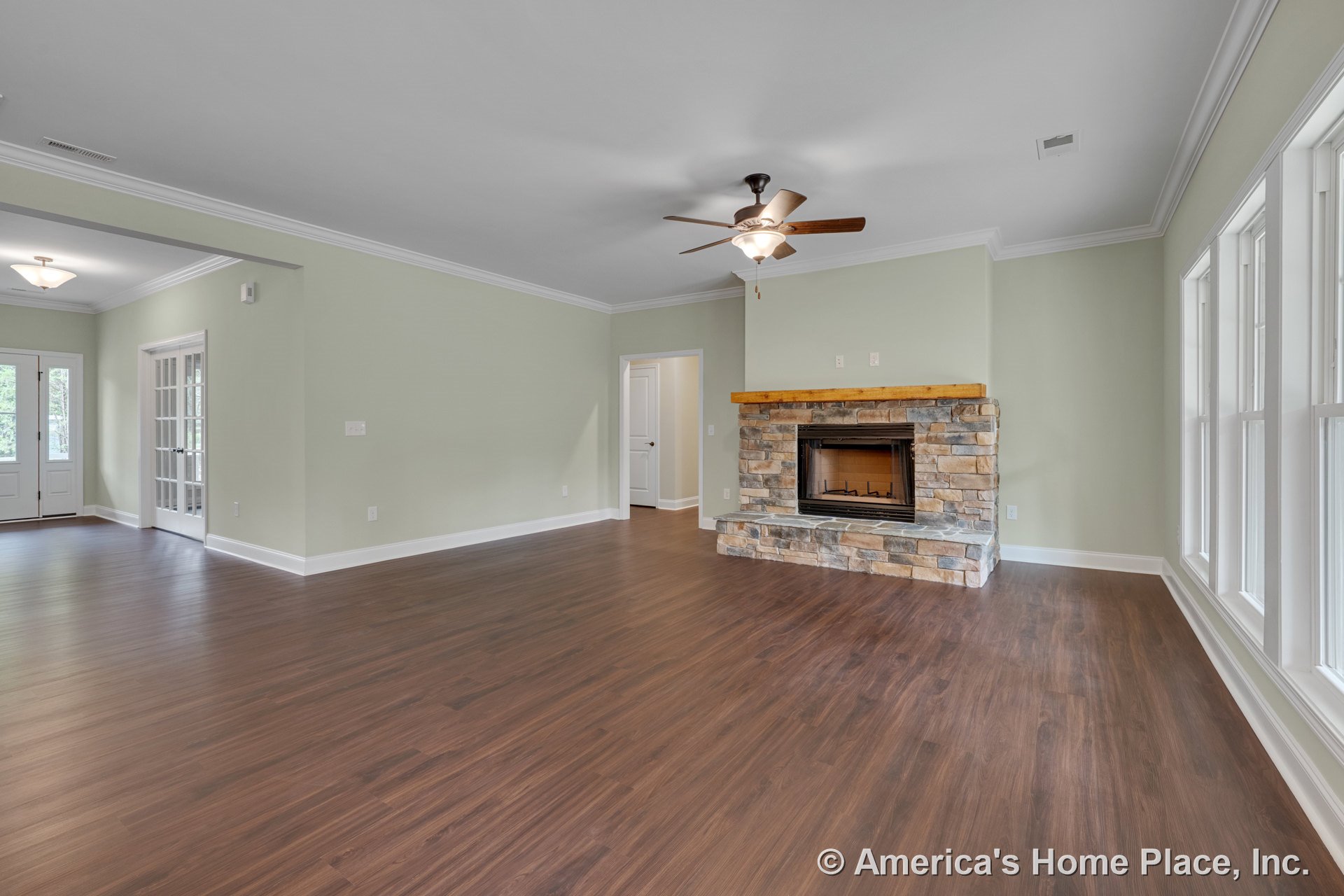 Stone fireplace with wood mantel, wide plank wood-look flooring, large windows framed in white trim, ceiling fan with light fixture, crown molding, and double French doors leading