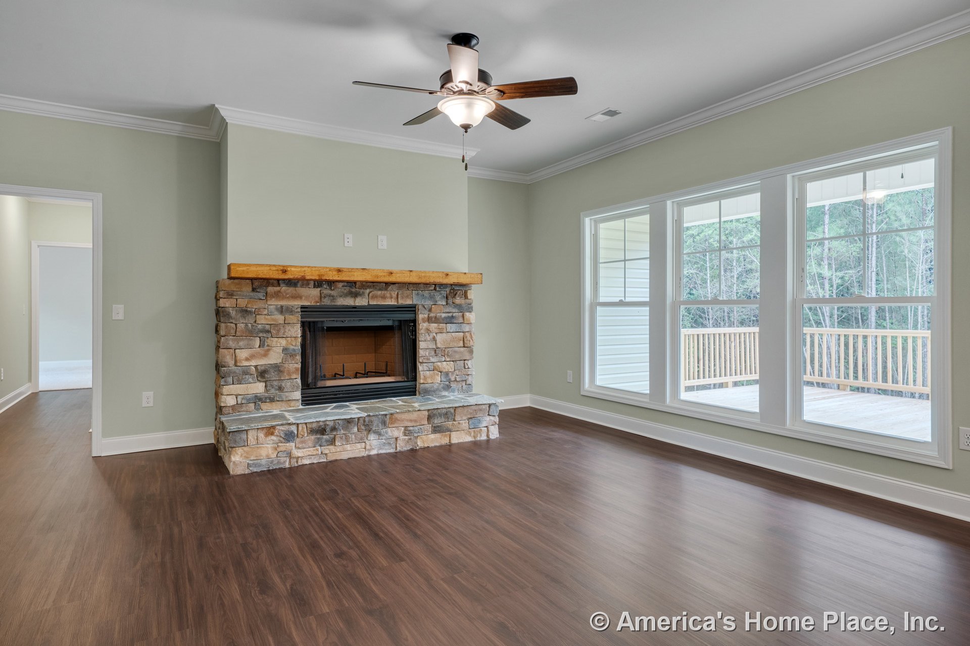 Stone fireplace with wood mantel, large triple windows facing porch, ceiling fan with light fixture, wood-look flooring, and crown molding trim in spacious living room.