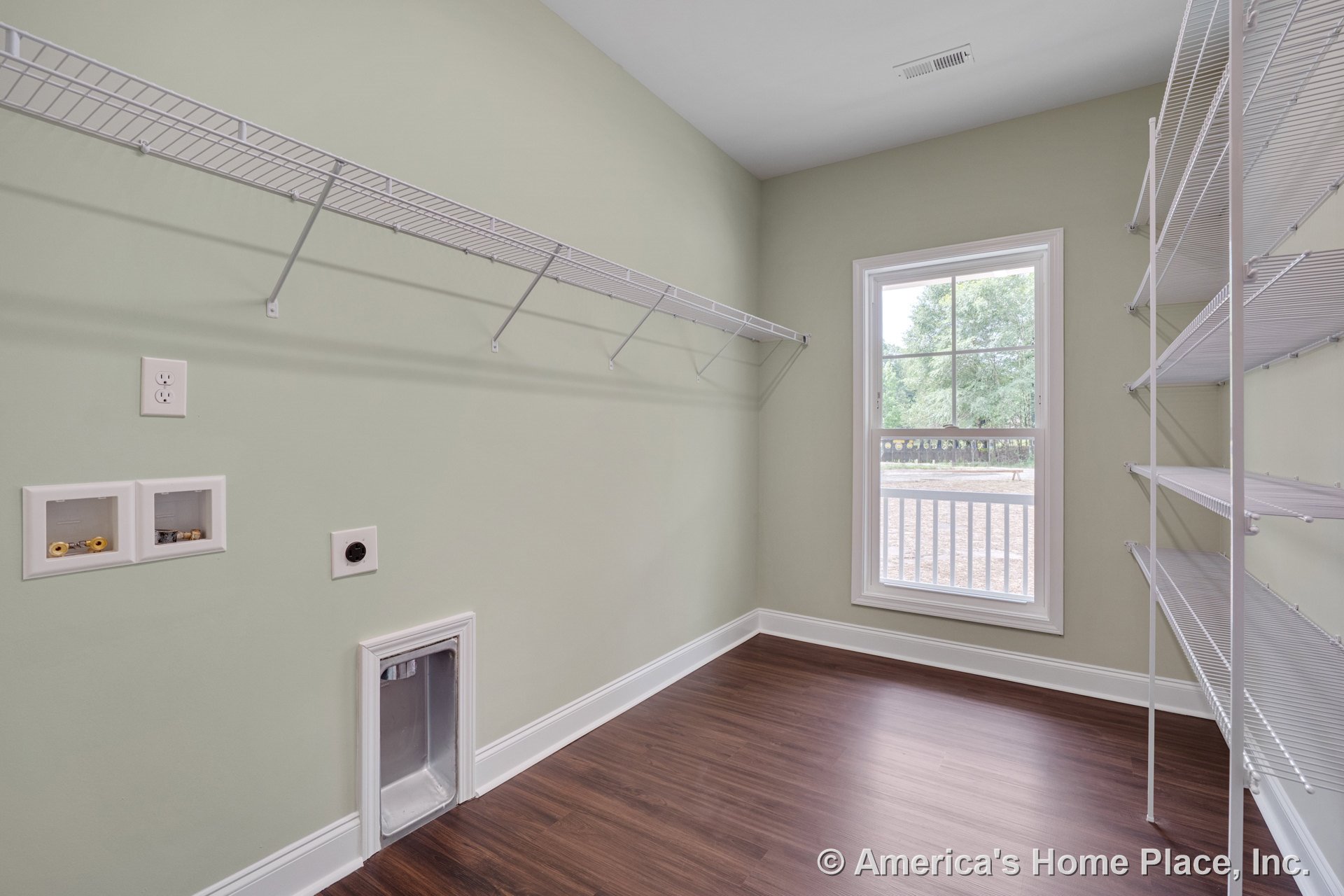 Laundry room with wire wall shelving, utility hookups, large window with white trim, vinyl plank flooring, baseboard molding, wall vent, electrical outlet, and light green walls.