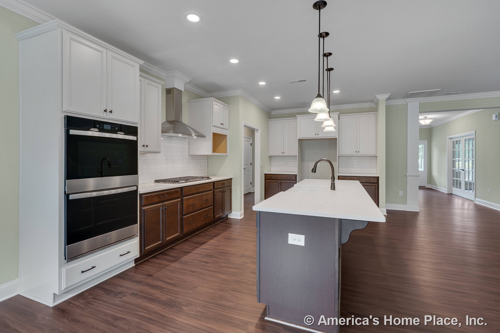 Spacious kitchen with large quartz island, double wall ovens, stainless steel range hood, pendant lights, white upper cabinets with crown molding, dark wood lower cabinets, subway