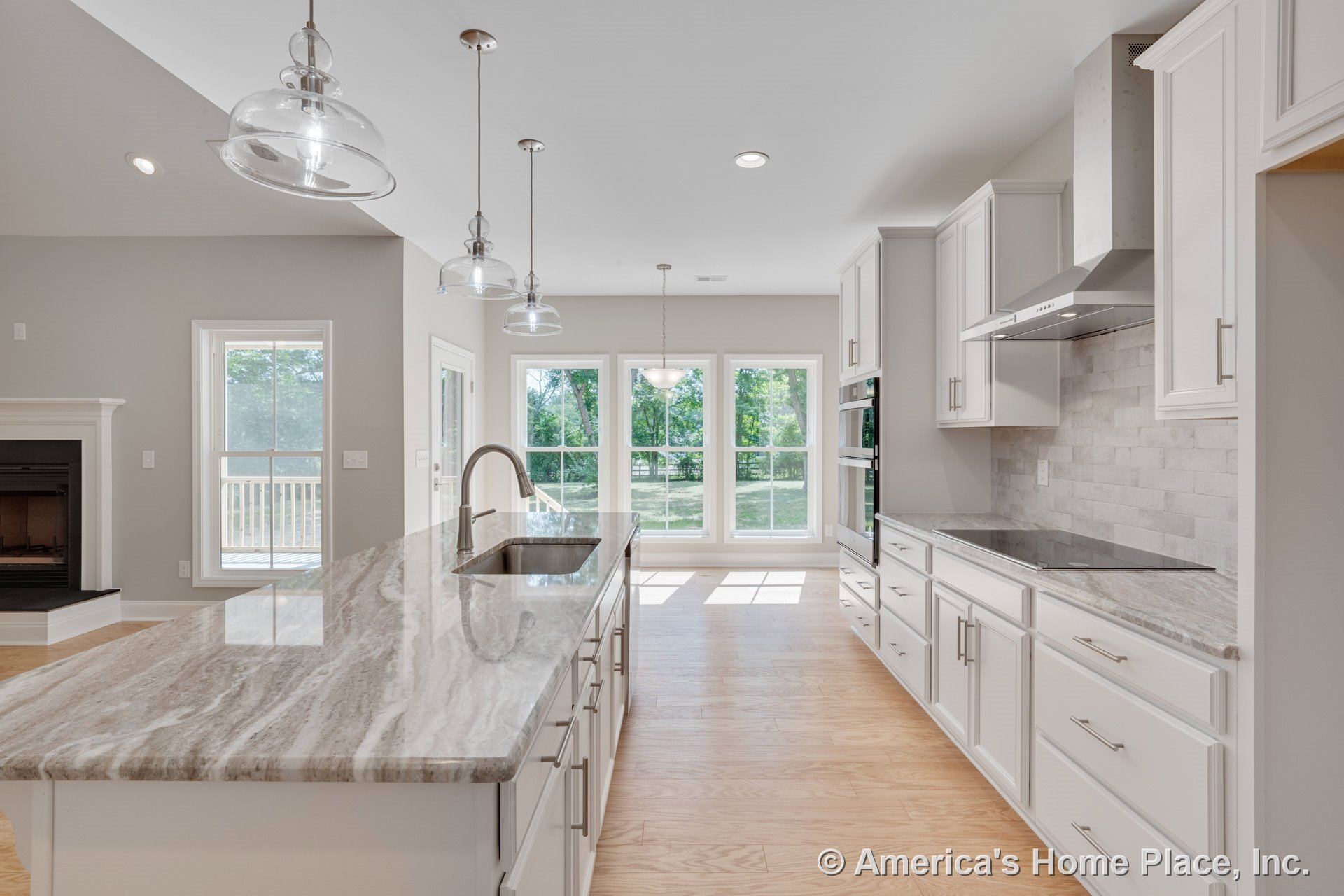 White kitchen with marble-look countertops, tile backsplash, and light wood flooring; large windows along the back wall; central island with undermount sink and glass pendant
