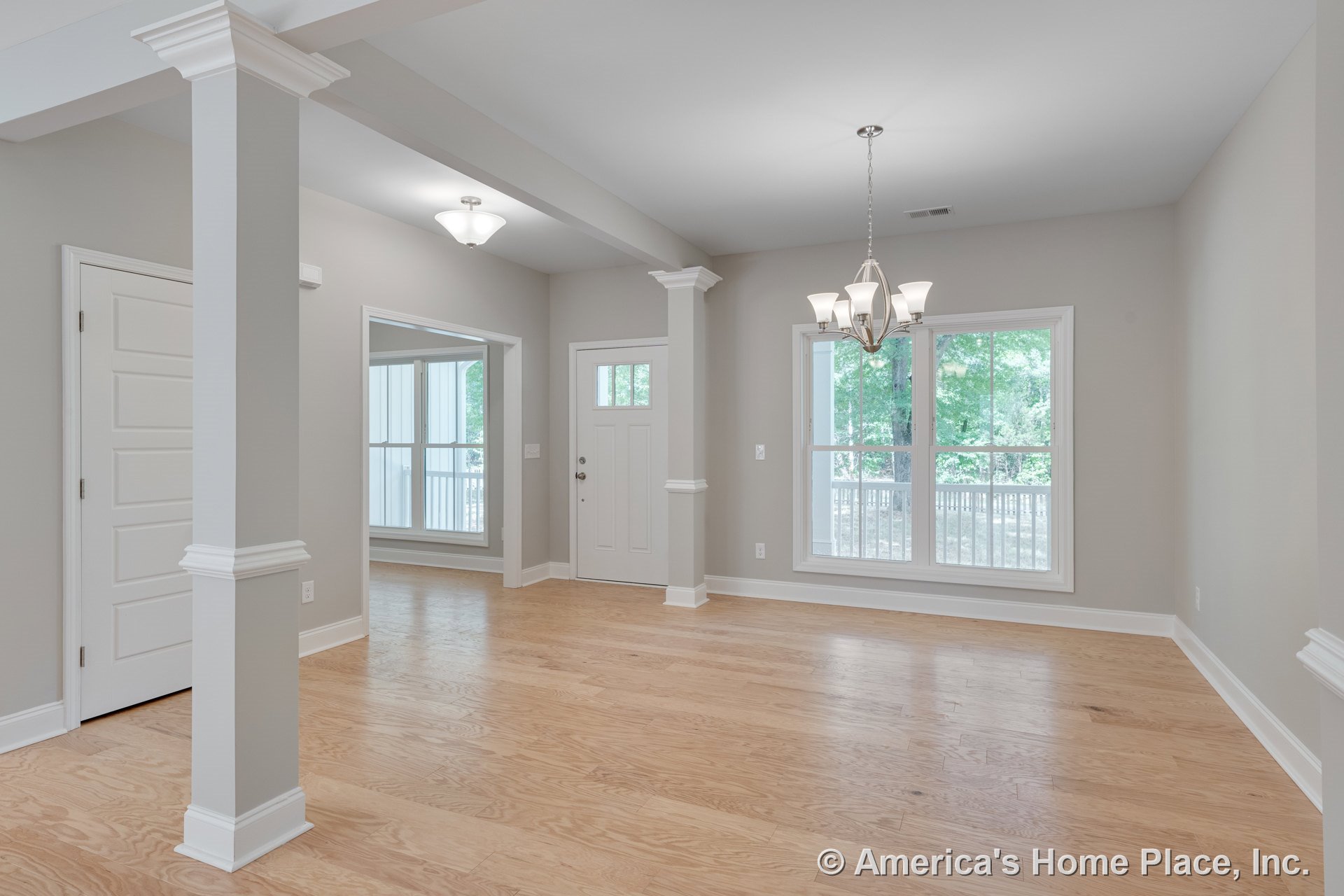 Dining area with light wood flooring, large double windows, modern chandelier, white trim and columns, entry door with transom window, and neutral wall paint in an open layout.