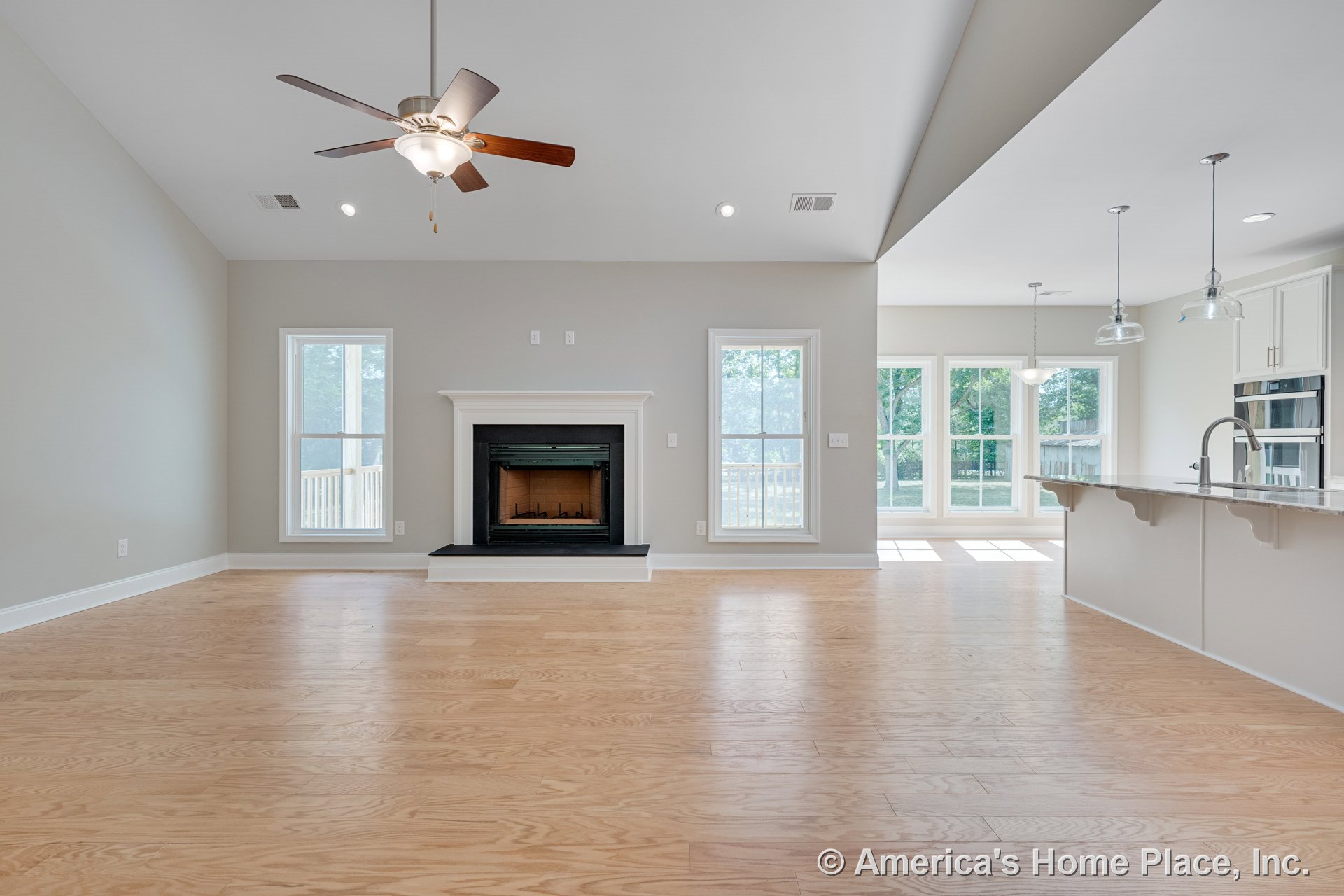 Central fireplace with white mantel, light wood flooring, large vertical windows, open kitchen with peninsula and cabinetry, pendant lights above countertop, ceiling fan with