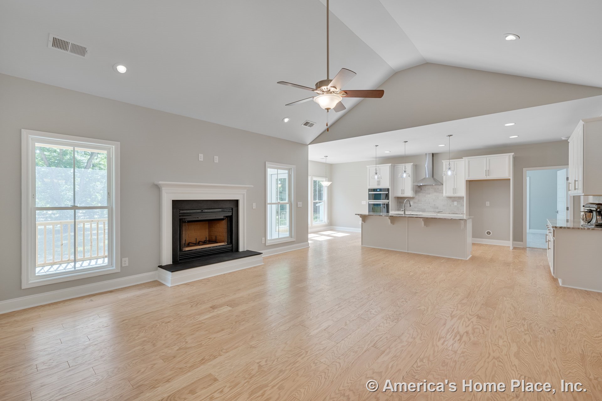 Fireplace with white mantel beneath vaulted ceiling with recessed lighting, large windows with trim, light wood flooring, kitchen island with pendant lights, white cabinetry, stone
