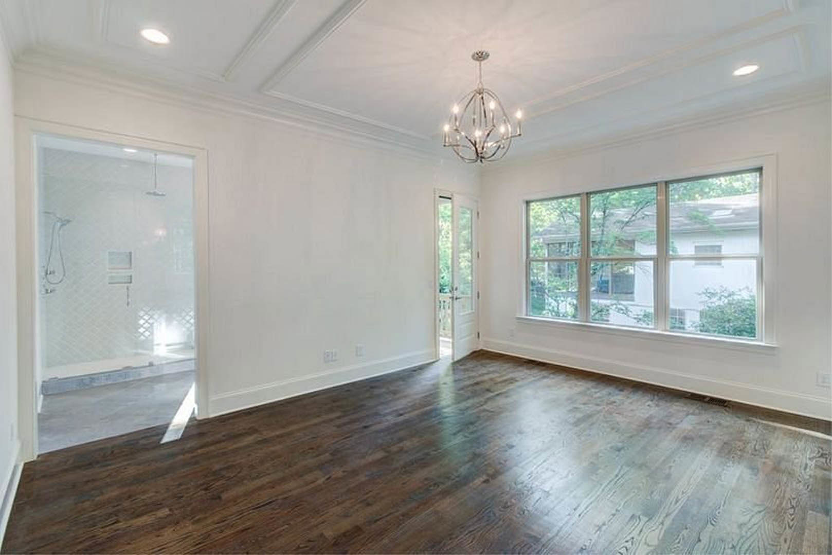 Chandelier hanging from white ceiling above dark hardwood floors, large window with view of trees, glass shower enclosure visible in background