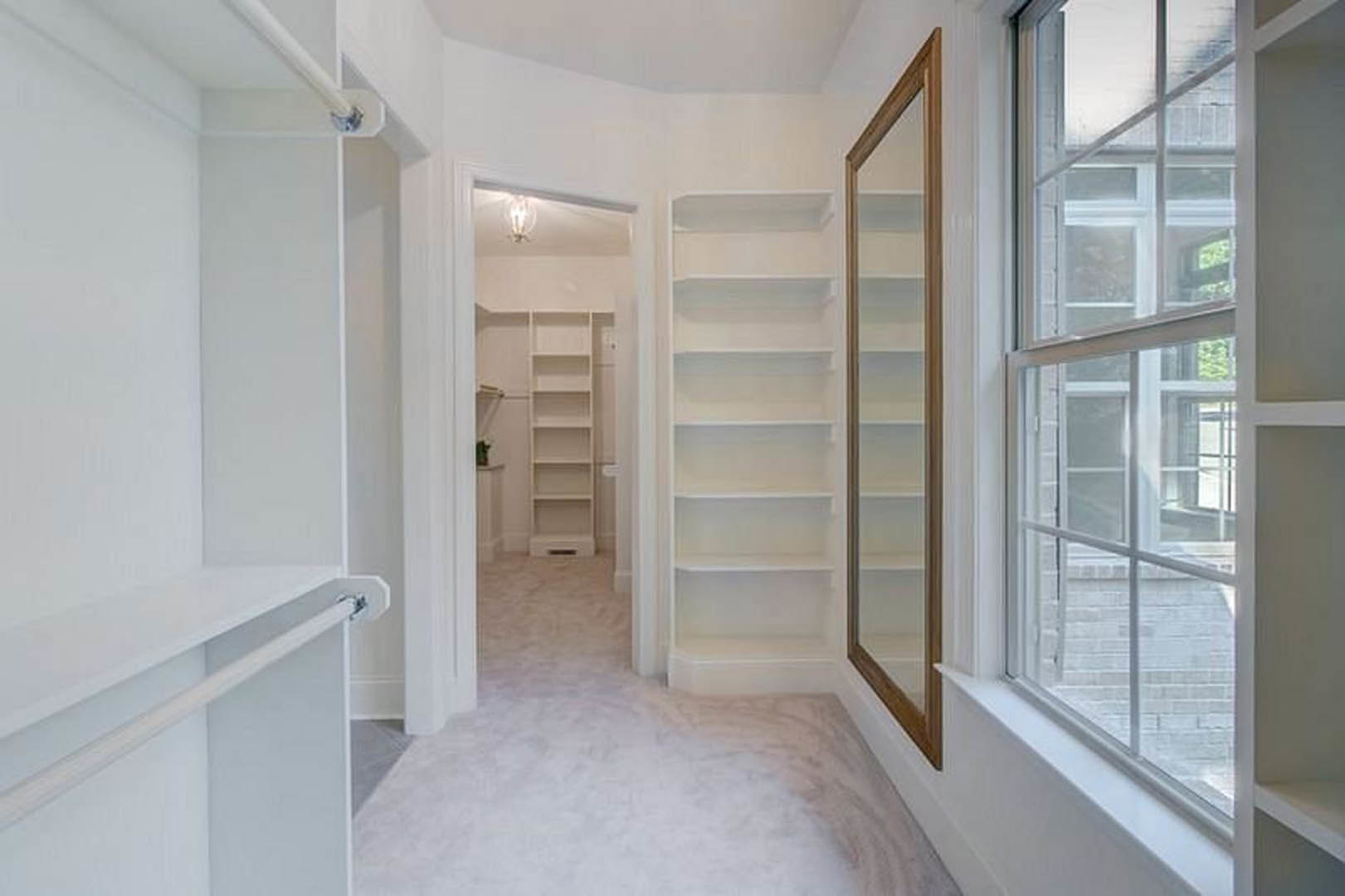 White-walled room featuring built-in white shelves, a window with a white frame, a close-up mirror, and a hanging rod.