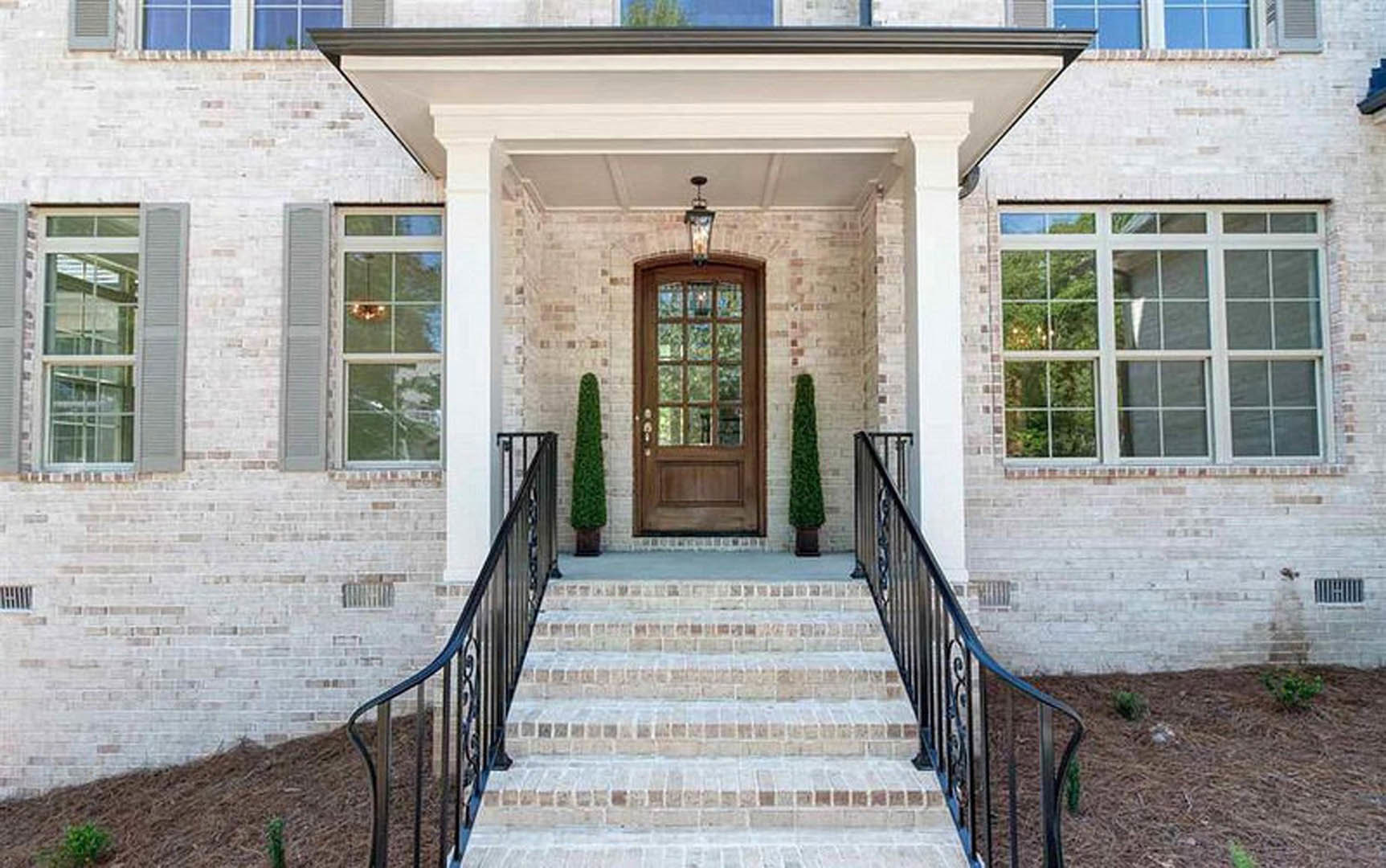 Wooden front door with glass panes, white trim, multi-pane window, brick steps, metal handrail, and green shrub along porch exterior