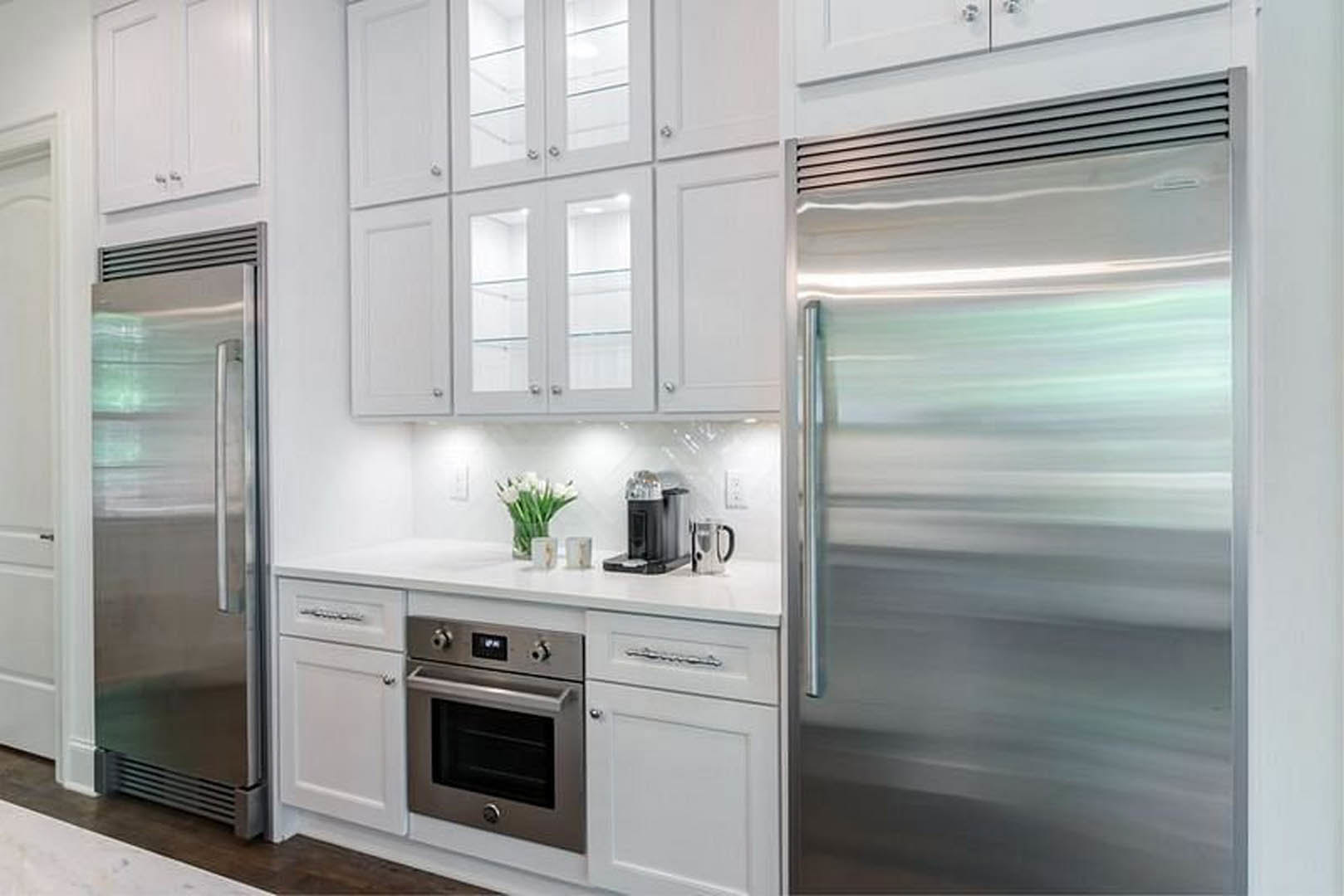 White cabinetry and quartz countertops frame stainless steel appliances, including a built-in oven and coffee machine; a glass door and a bouquet of white flowers in a clear vase