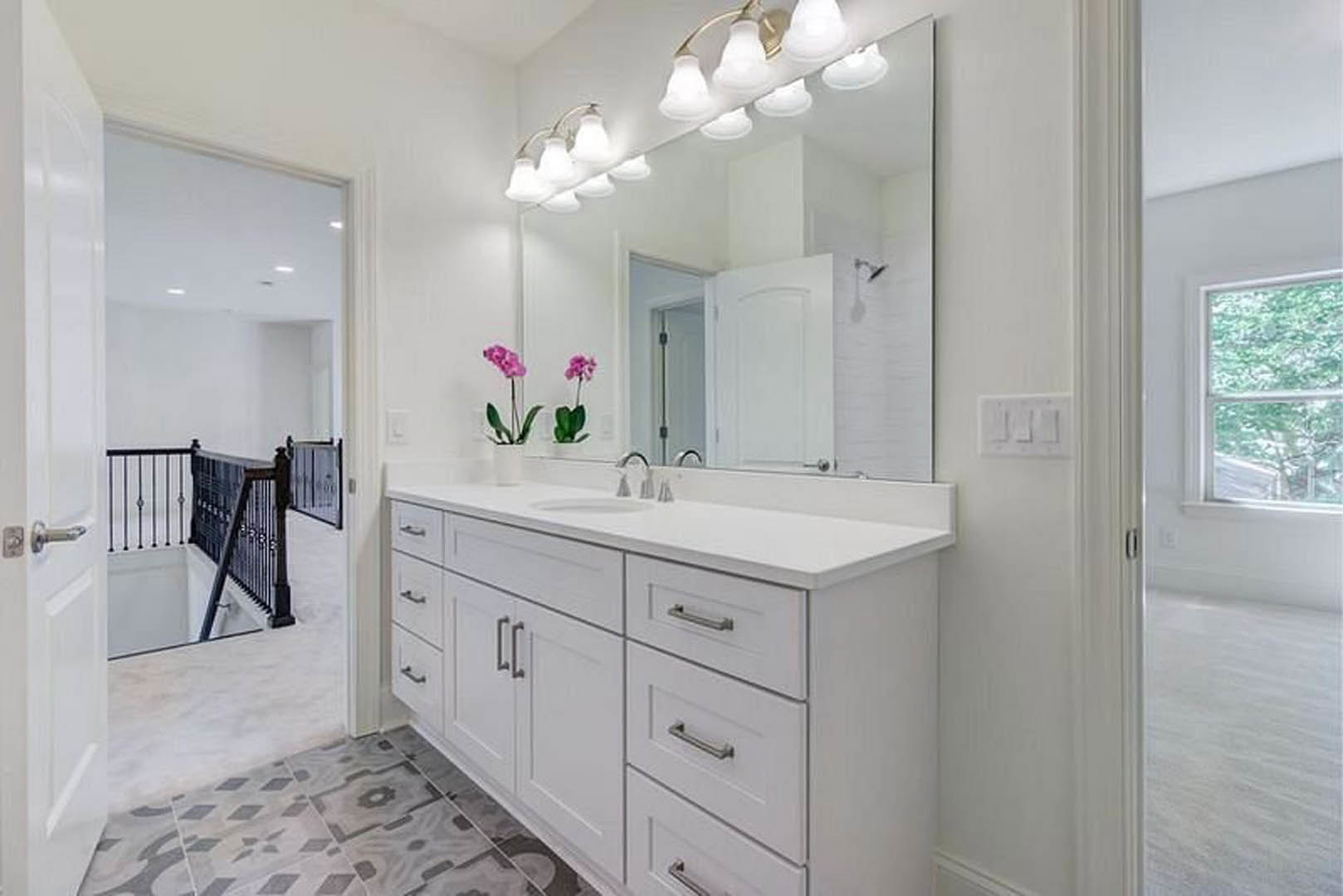 Bathroom with tiled walls, wide mirror above a stone countertop and sink, row of lights mounted on mirror, window overlooking trees, cabinetry with drawers, chrome tap, close-up of