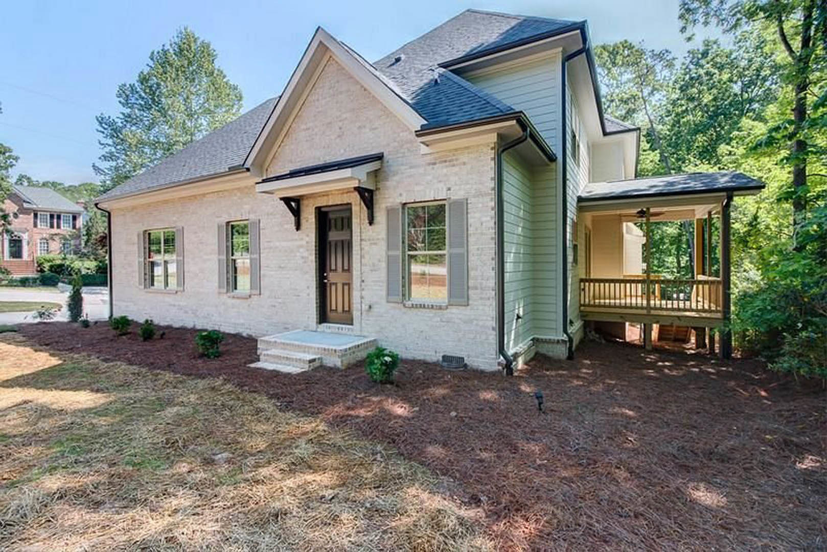 Two-story home with brick exterior, covered front porch, wooden deck, large windows, and landscaped yard with trees and plants
