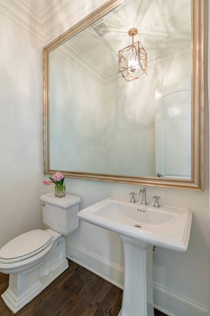 White tile bathroom featuring a rectangular mirror above a modern sink with chrome faucet, toilet with closed lid, pink flowers in a glass vase on the countertop, and a
