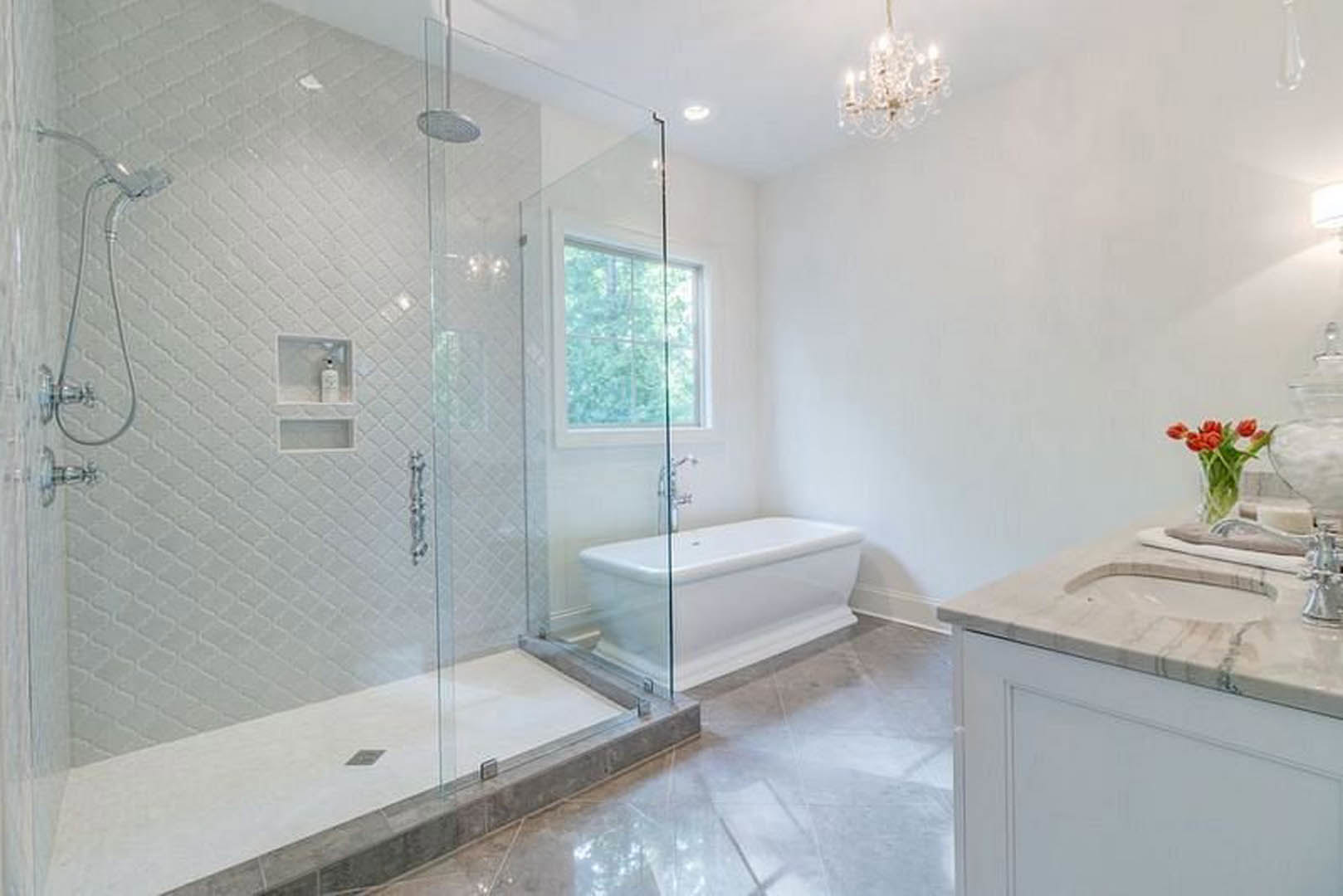 Bathroom with glass shower enclosure, freestanding white bathtub, white tile walls, red tulips in a glass vase on countertop, window with curtain rod, green plant in background