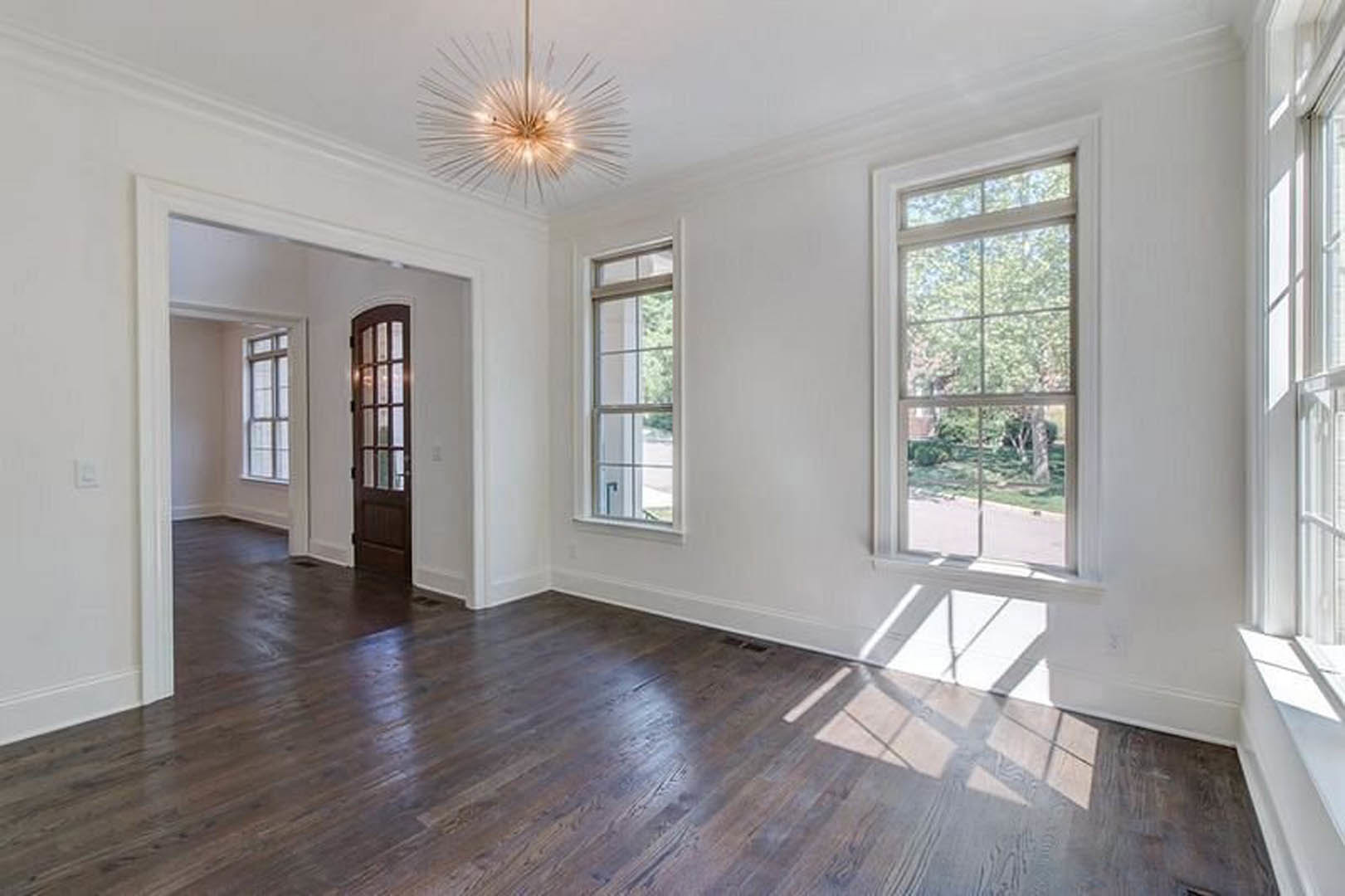 Wood flooring in a spacious room with a sparkling chandelier, large multi-pane window, and white wall molding.