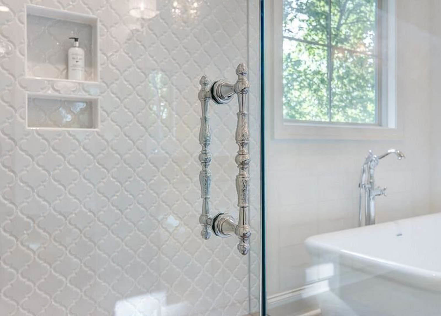 Glass door separating shower area, white freestanding bathtub, chrome faucet, candlesticks on tiled countertop, window with view of trees, white container, bathroom sink and mirror