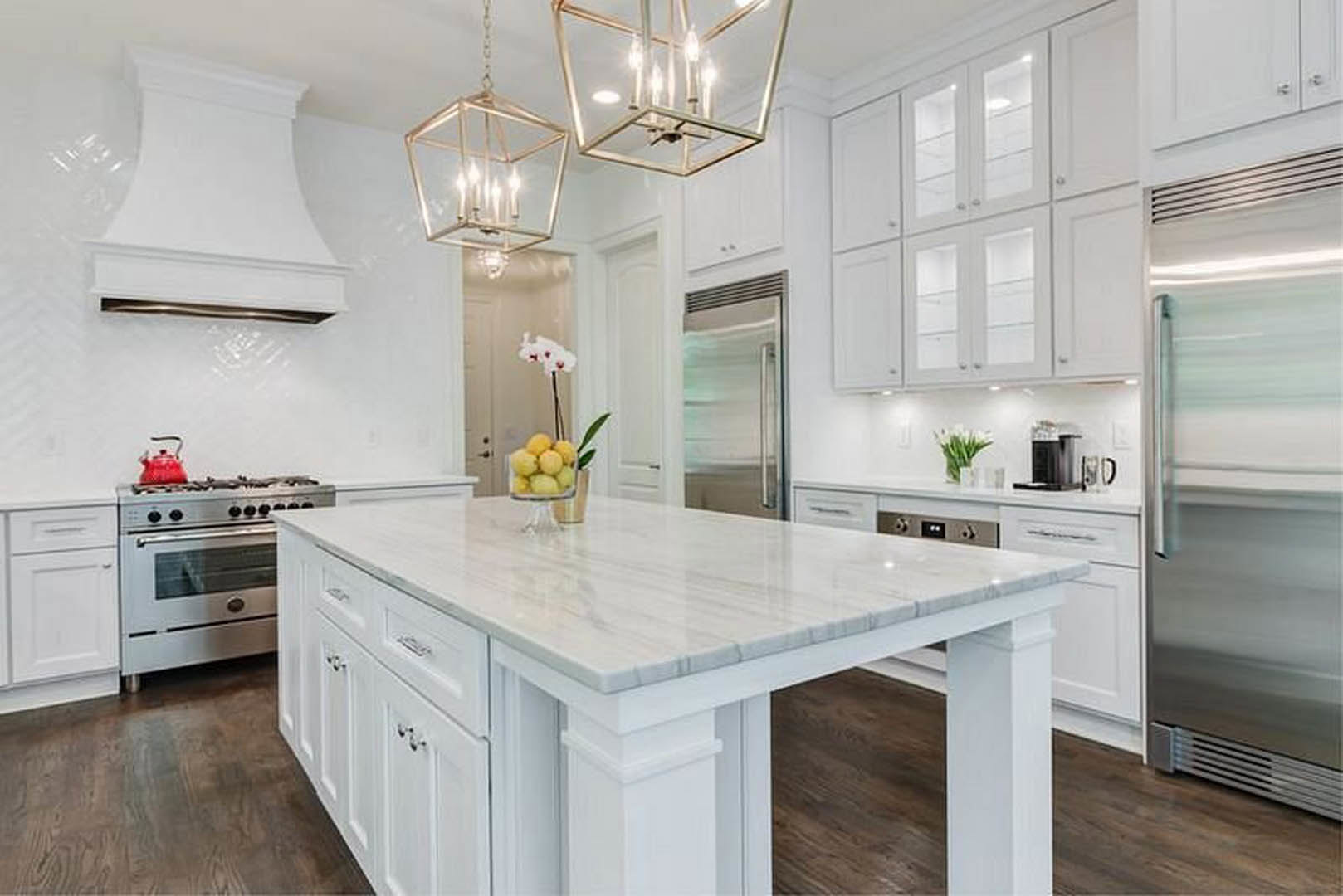 White kitchen with marble island, white cabinetry, bowl of lemons on countertop, stainless steel stove with black knobs, chandelier hanging from ceiling, window in background.