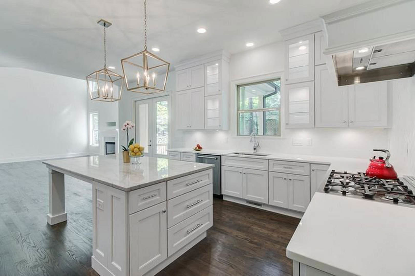 White kitchen with shaker cabinets, marble island countertop, stainless sink, red tea kettle on stove, pendant chandelier, hardwood floors