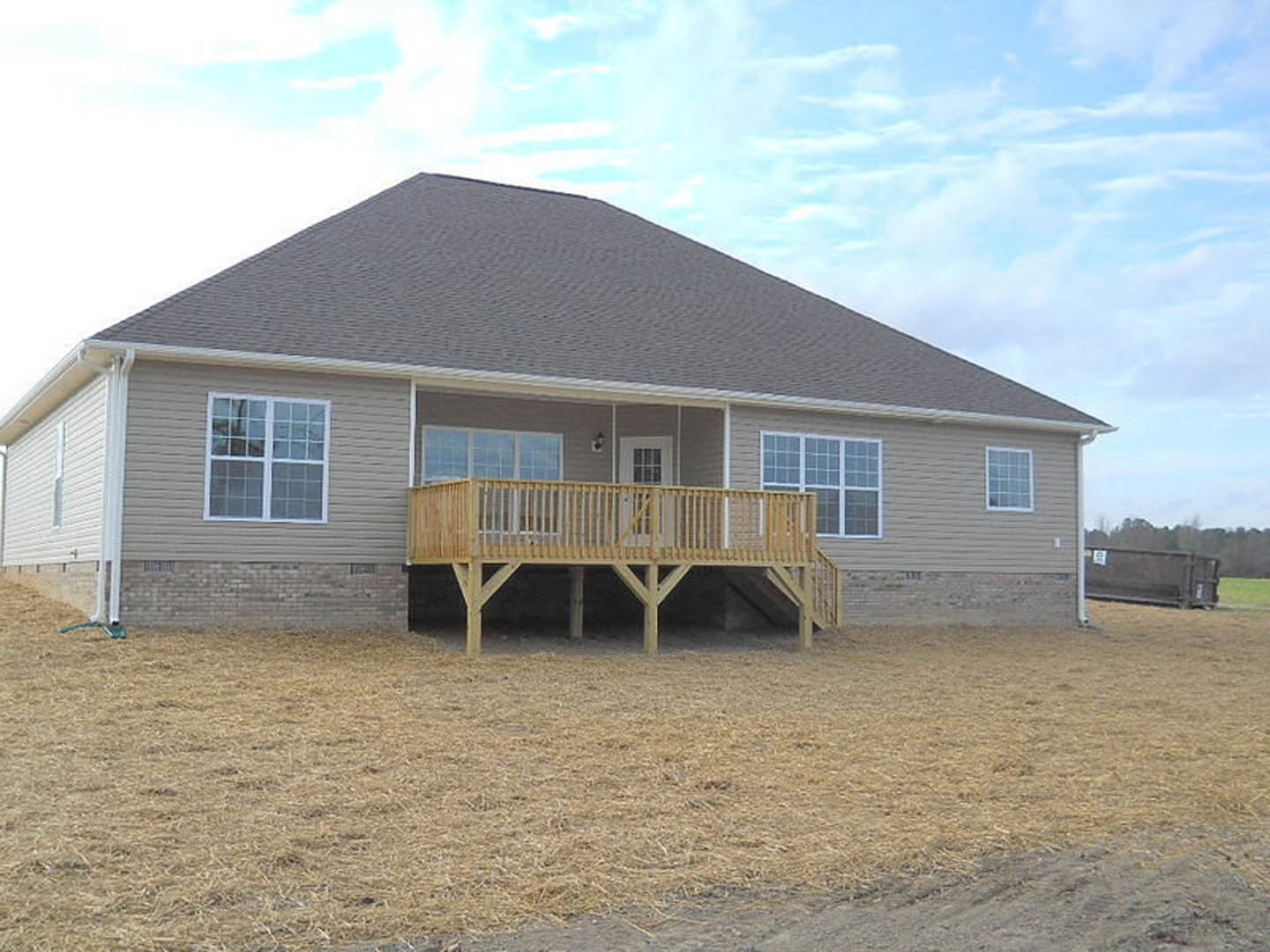 Wooden deck with railing overlooking grassy yard, multi-pane window on cottage-style home, hay scattered on ground, partly cloudy sky above