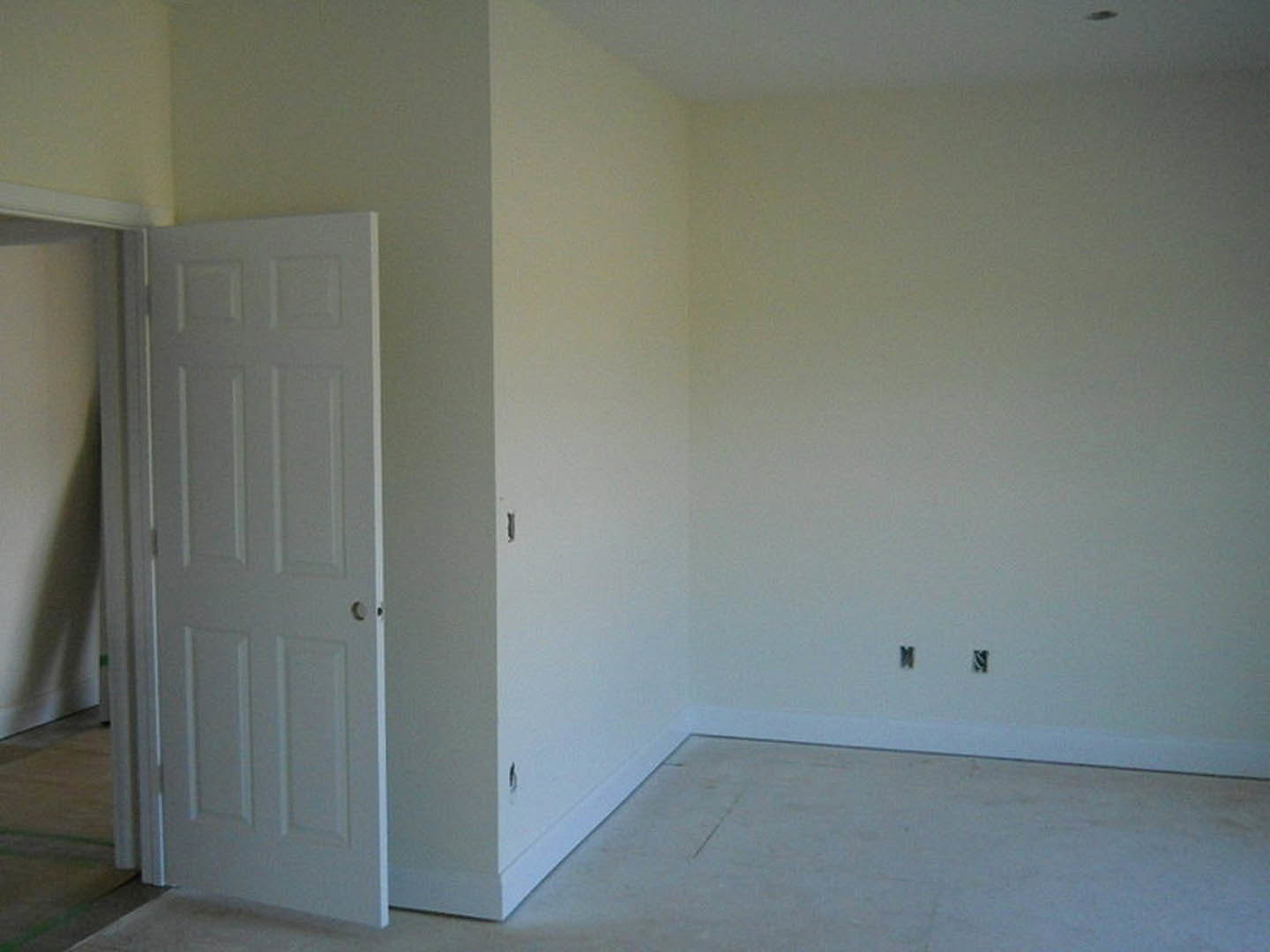 White paneled door with round hole, opening into a room with light-colored plaster walls and wood flooring; corner of a white wall and small table visible in foreground.