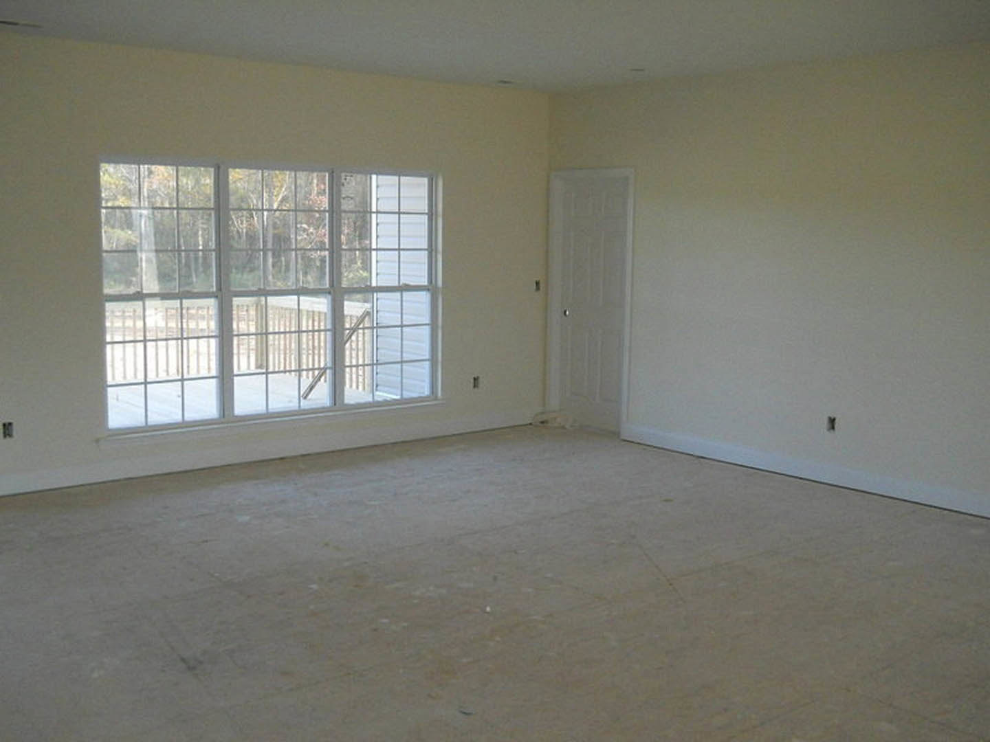Spacious room featuring light wood flooring, white plaster walls, a large window overlooking a deck with trees, and a white door with a silver knob
