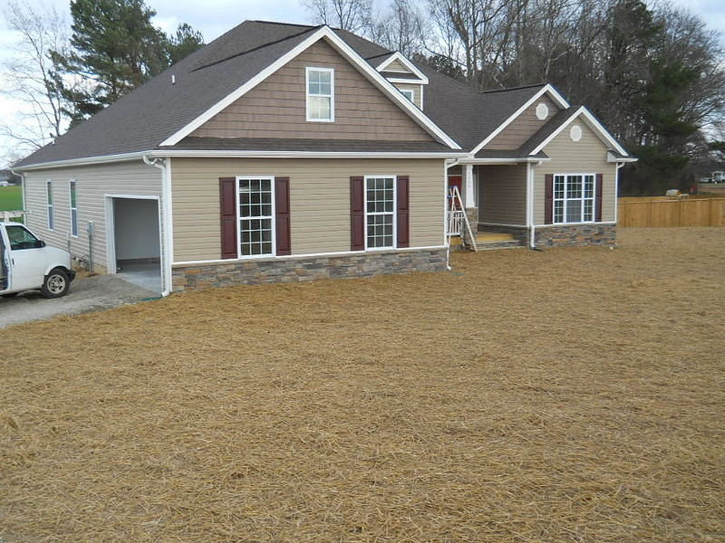 Two-story house with gray roof, multiple white-framed windows, attached garage, white pickup truck parked in driveway, green grass yard, and mature trees in background