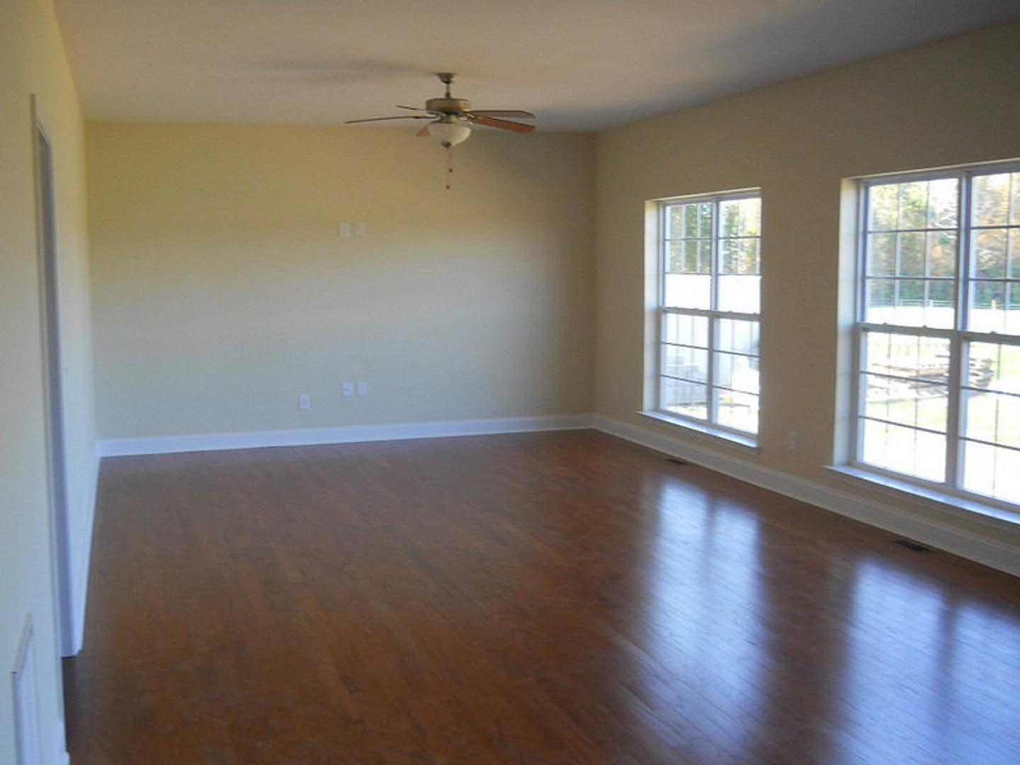 Ceiling fan with light fixture above wood laminate flooring, two windows with multiple panes letting in daylight, neutral walls, residential interior.