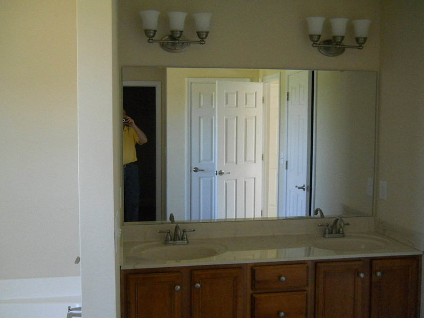 Large bathroom mirror above a white countertop with multiple cabinet doors, chrome faucet, and three-light fixture; blurred reflections of a person and bowl visible.