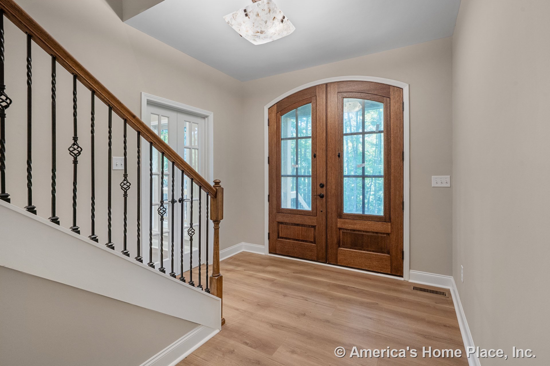 Arched double wooden entry doors with glass panels, light wood flooring, staircase featuring wood railing and wrought iron balusters, modern ceiling light fixture, neutral wall