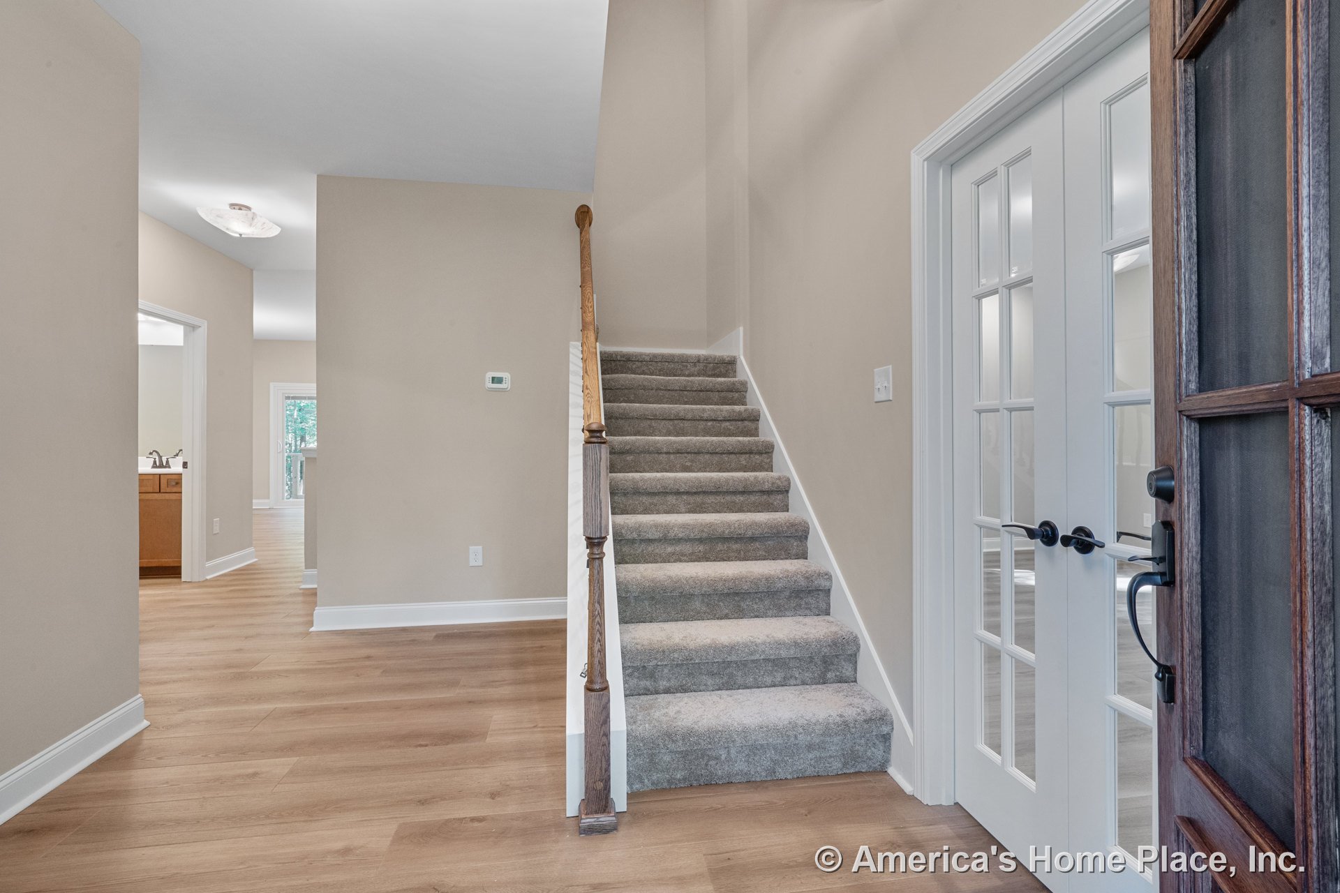 Carpeted staircase with wood handrail, light wood flooring, double glass French doors, white baseboards, neutral wall color, and ceiling lighting fixture in the entryway.