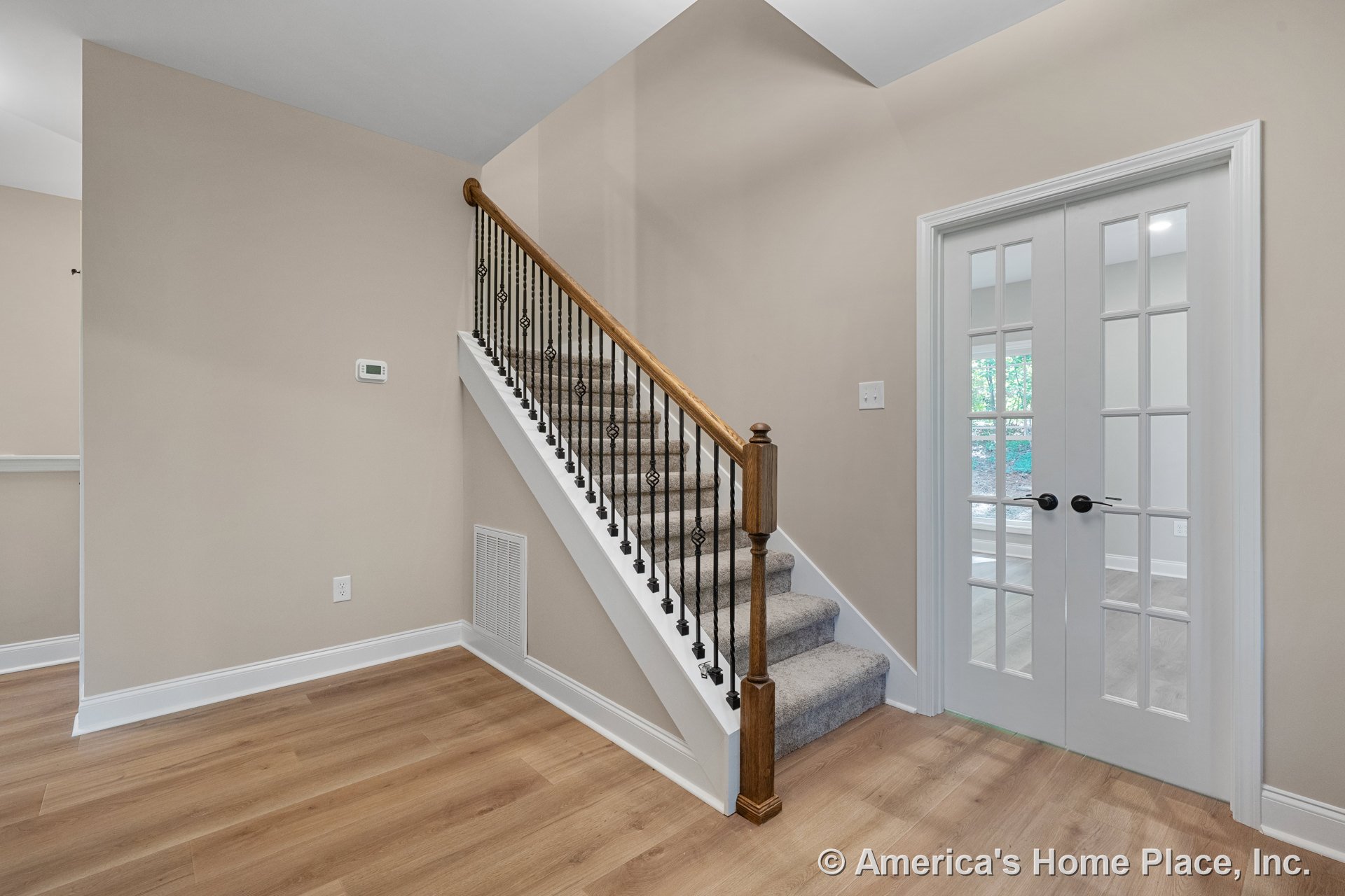 Carpeted staircase with iron balusters and wood handrail beside double French doors with glass panels, light wood flooring, white baseboards, and neutral wall paint in foyer.