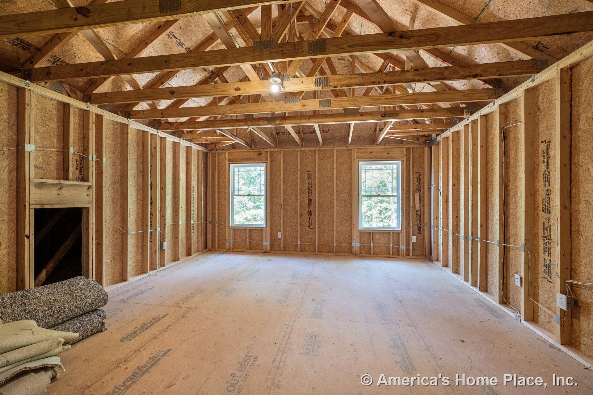 Unfinished room with exposed wood framing, plywood subfloor, vaulted ceiling supported by trusses, two double-hung windows, visible electrical wiring, and a single ceiling light