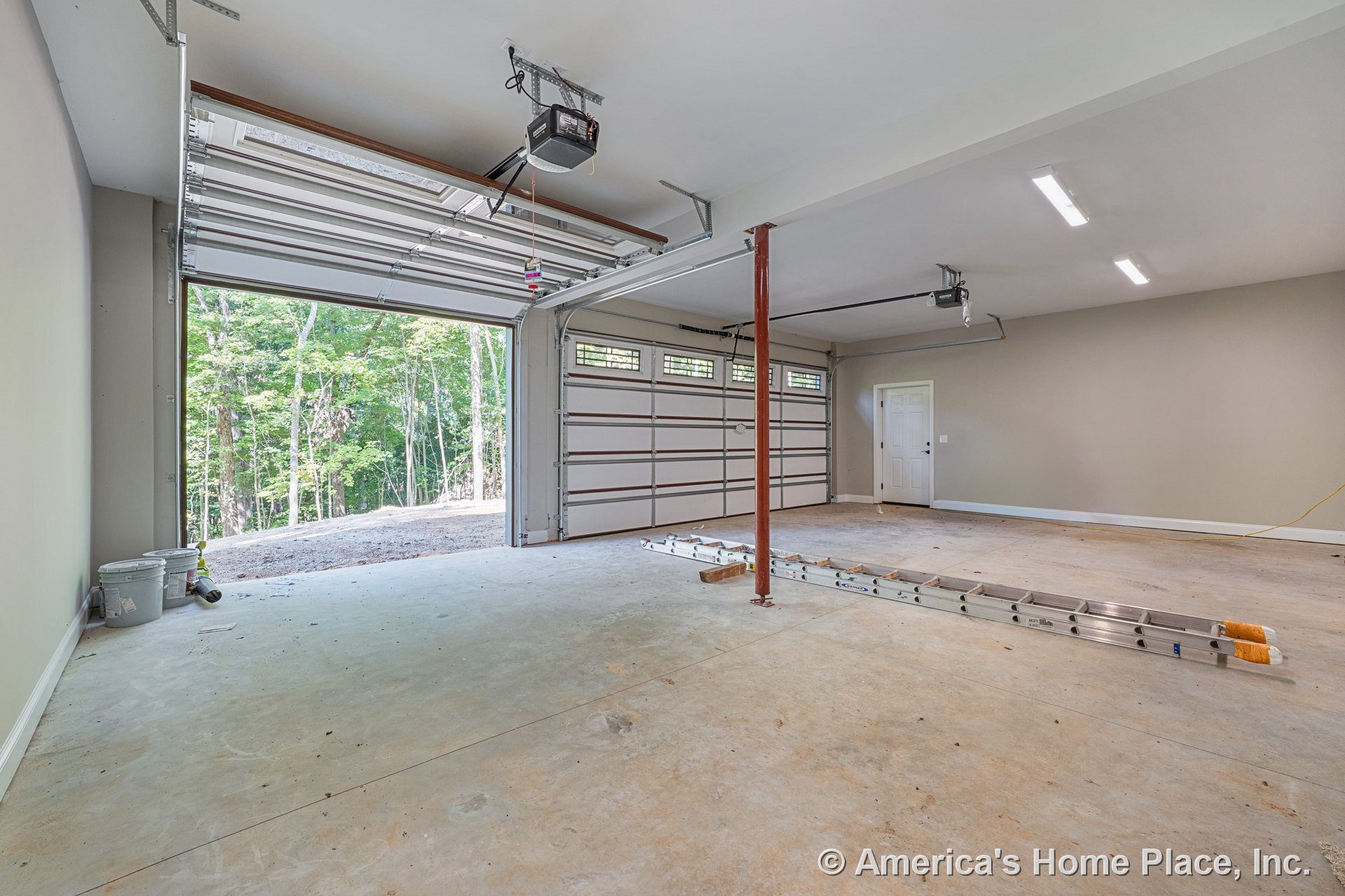 Two overhead garage doors with upper windows, concrete slab floor, ceiling-mounted fluorescent lights, interior entry door, exposed structural support column, finished walls, and