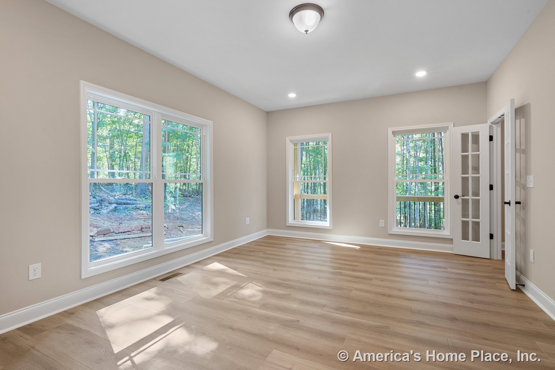 Large double-hung windows and a French door with glass panels allow natural light into a bedroom with light wood plank flooring, neutral wall paint, white baseboard trim, and a