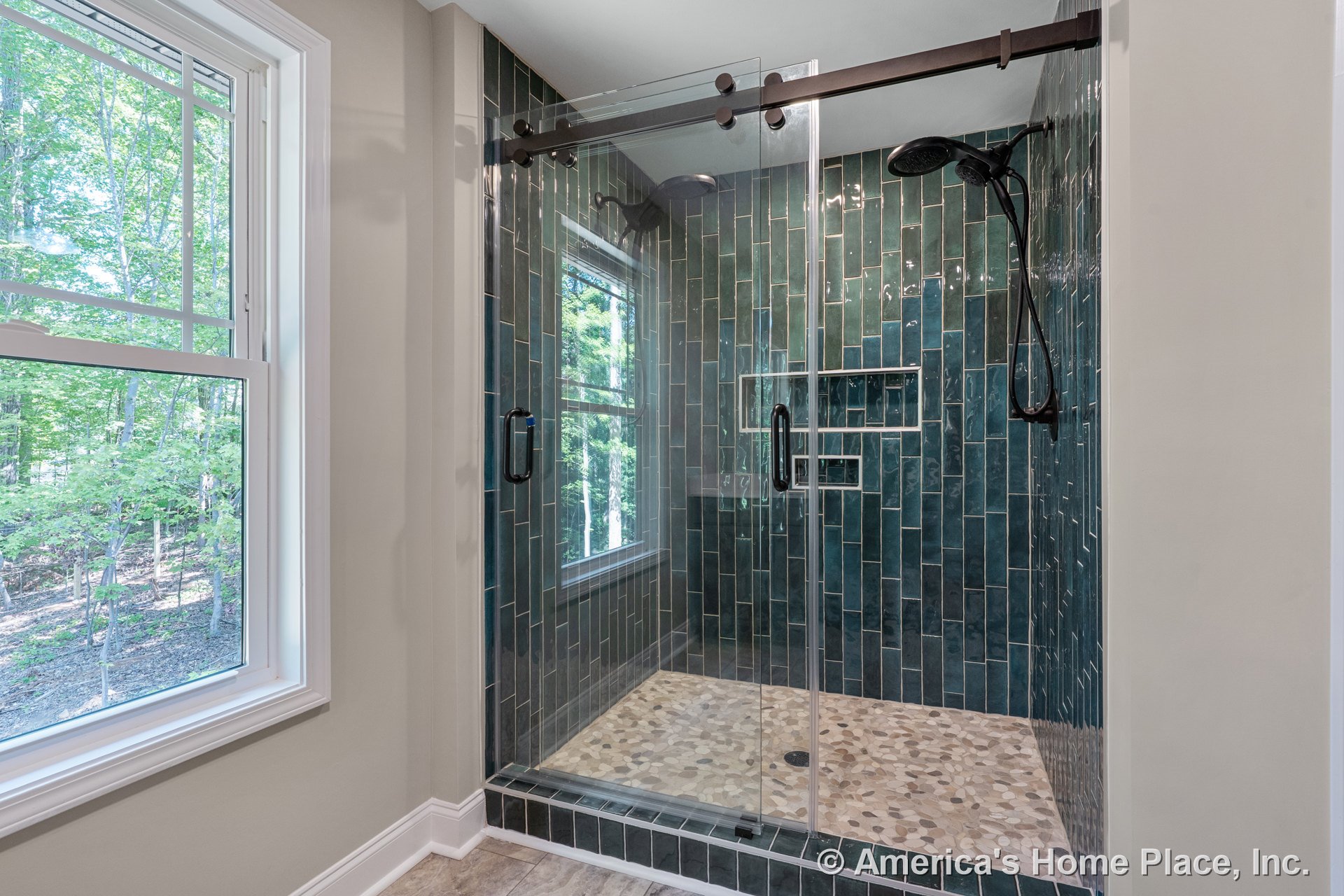 Walk-in shower with green vertical tile walls, frameless glass door, built-in shower niche, pebble stone floor, and large window with white trim.