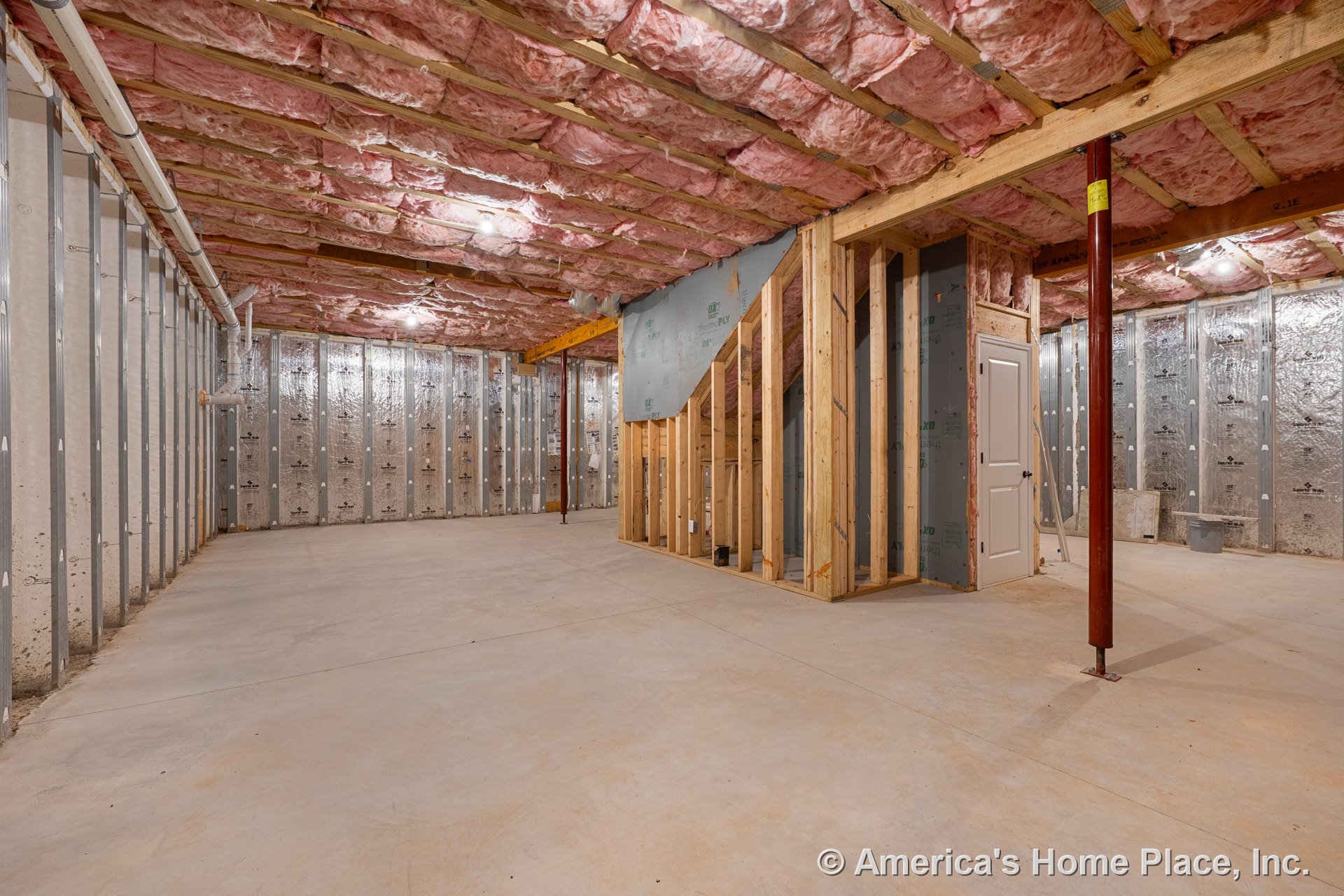 Unfinished basement with exposed ceiling insulation, concrete slab flooring, metal wall studs, framed staircase, visible utility pipes, single interior door, support column, and