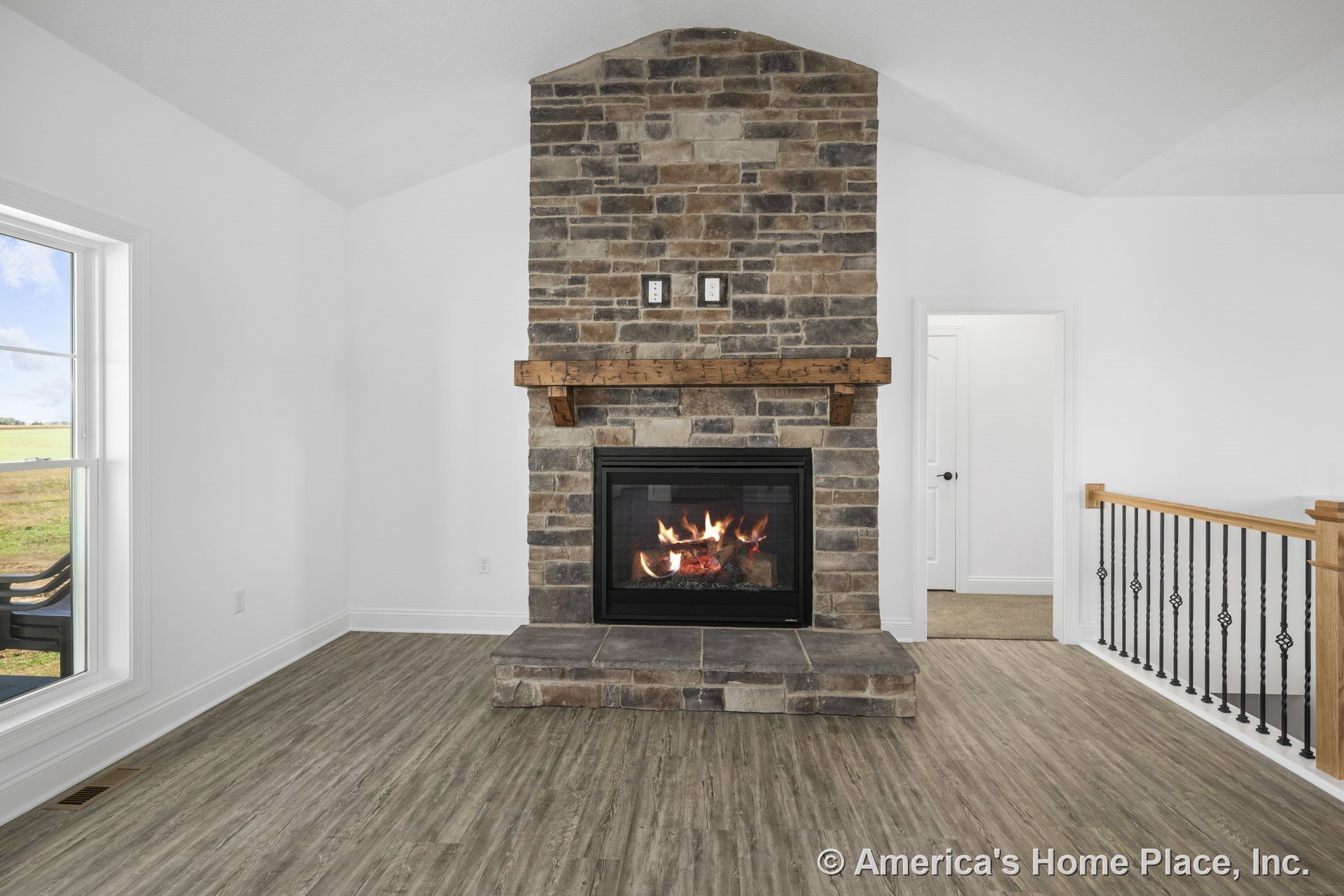 Stone fireplace with wood mantel, vinyl plank flooring, white walls and trim, large divided-light window, iron and wood stair railing, open layout.