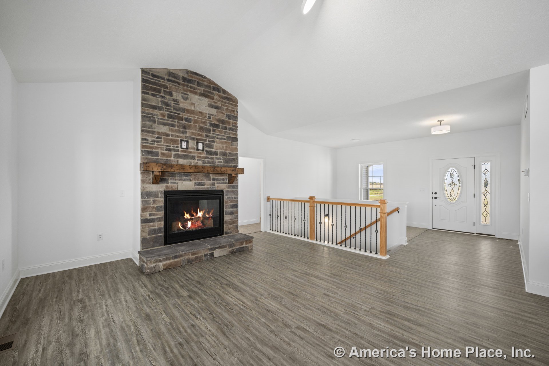 Stone fireplace with wood mantel, vinyl plank flooring, white trim and walls, decorative glass front door, open stair railing with wood and iron balusters, ceiling light fixture