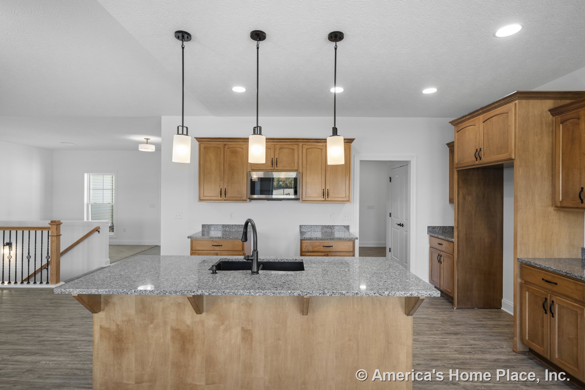 Granite kitchen island with wood upper and lower cabinets, built-in microwave, pendant lights, stainless sink with black faucet, open stair railing, recessed lighting, and hard
