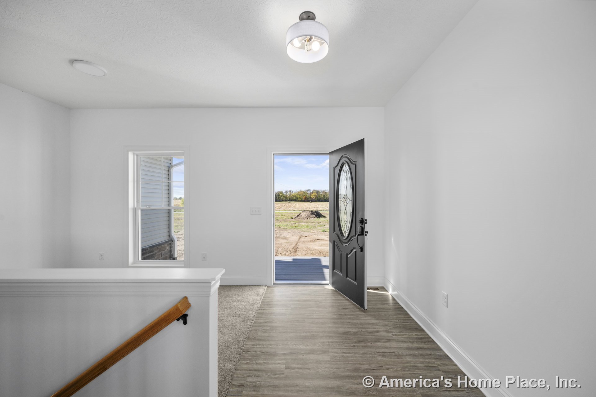 Black front door with decorative glass, vinyl plank flooring, white painted trim, stair railing with wood accent, flush mount ceiling light, large window filling foyer with natural