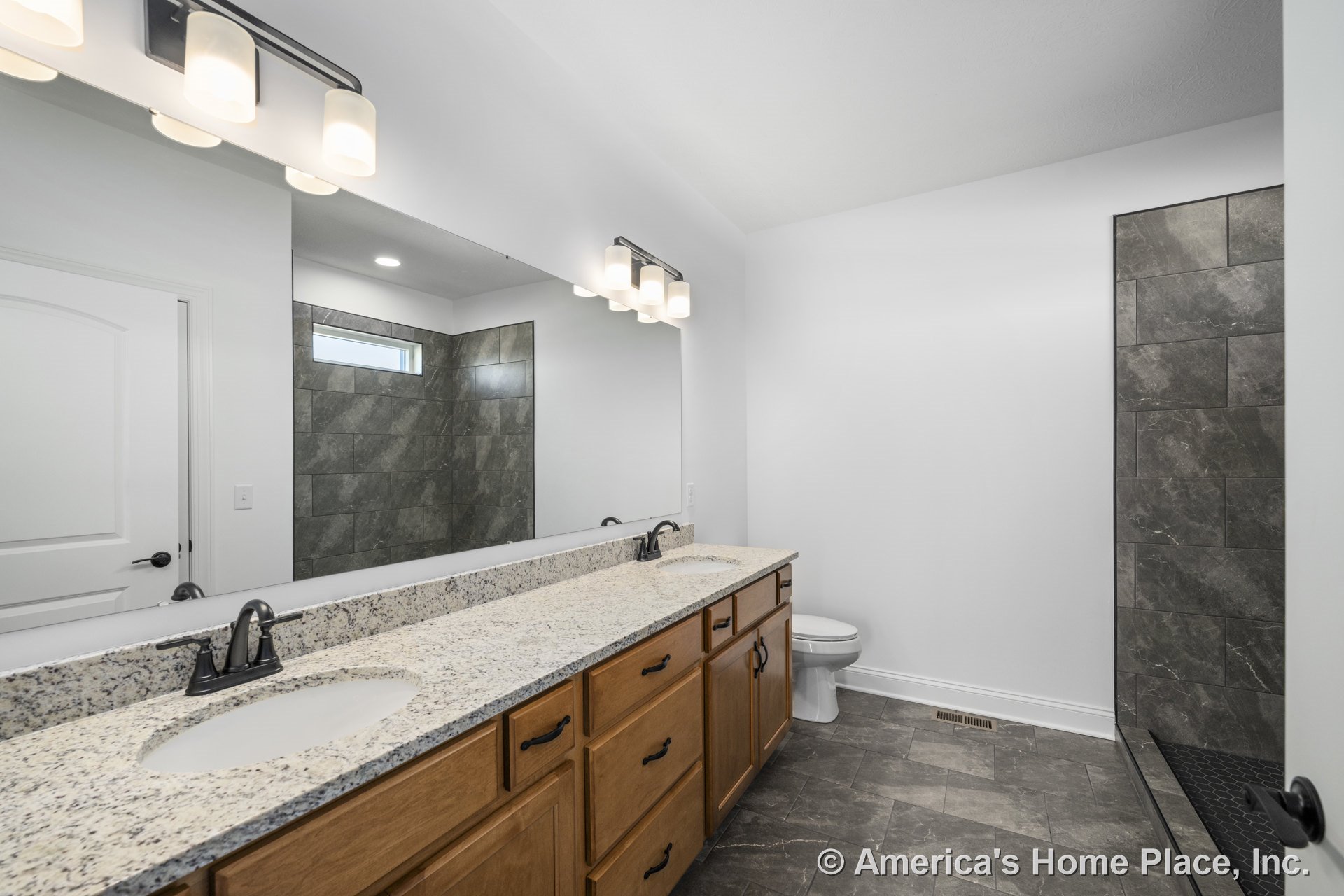 Double vanity with granite countertop and wood cabinetry, large wall mirror above, walk-in tiled shower with tile flooring, built-in overhead lighting, black hardware, white walls